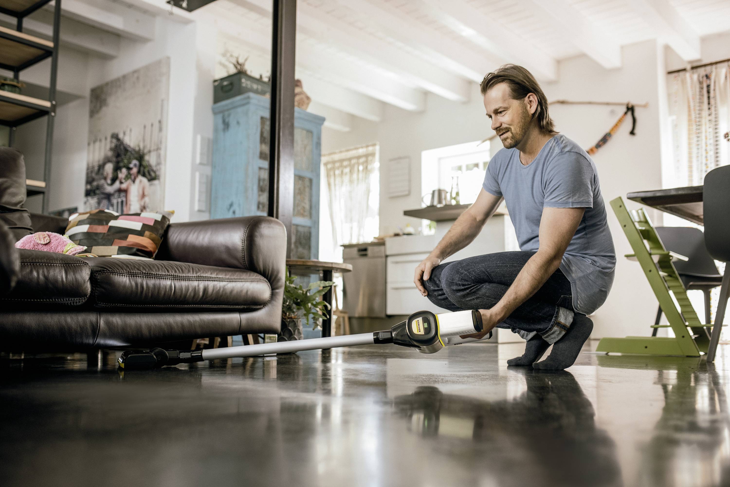 A man is hoovering the floor in a living room with a cordless vacuum cleaner. A sofa and kitchen furnishings are visible in the background.