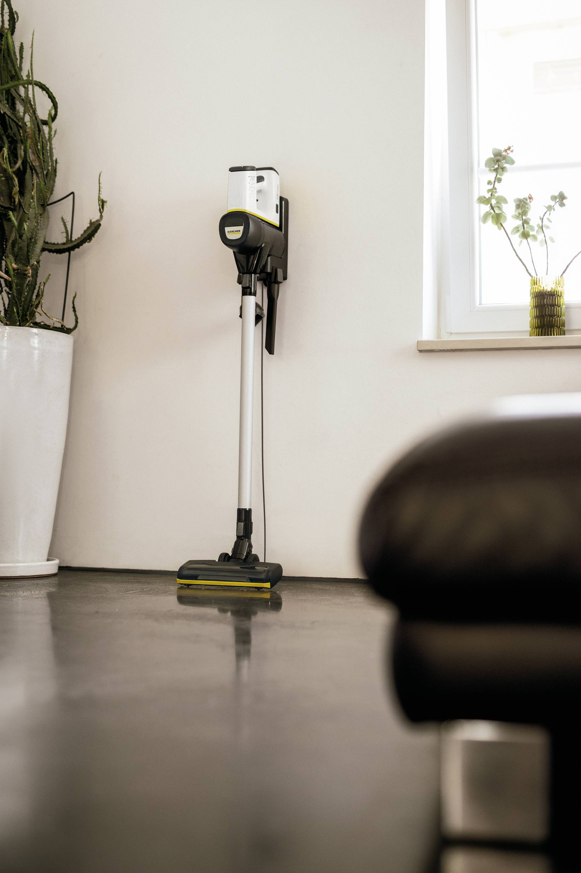 Cordless vacuum cleaner leaning against a white wall, next to a large plant pot, in a modern, well-lit room.