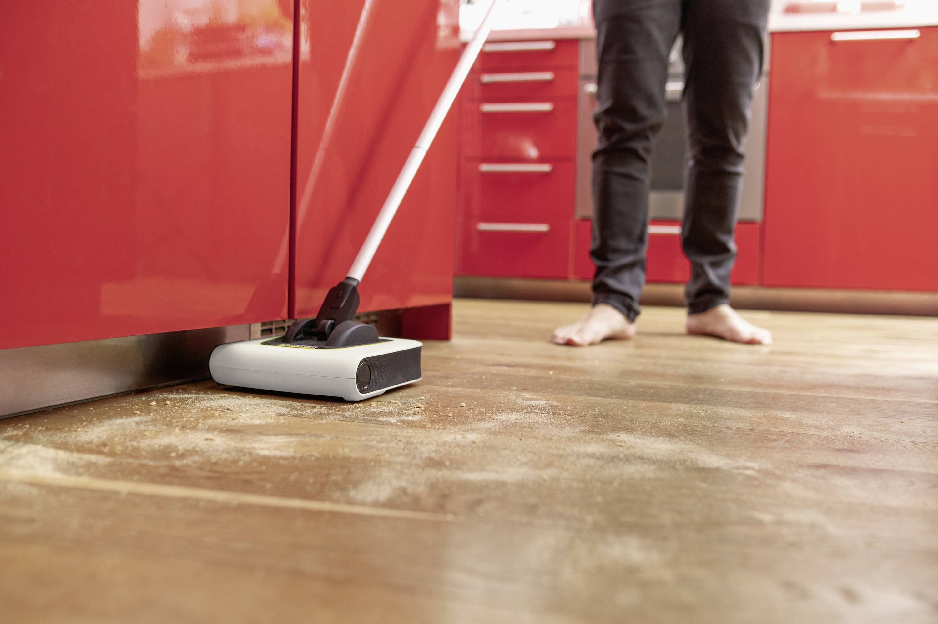 A person is mopping the wooden floor barefoot in a red kitchen with a floor mop, removing dust and dirt.