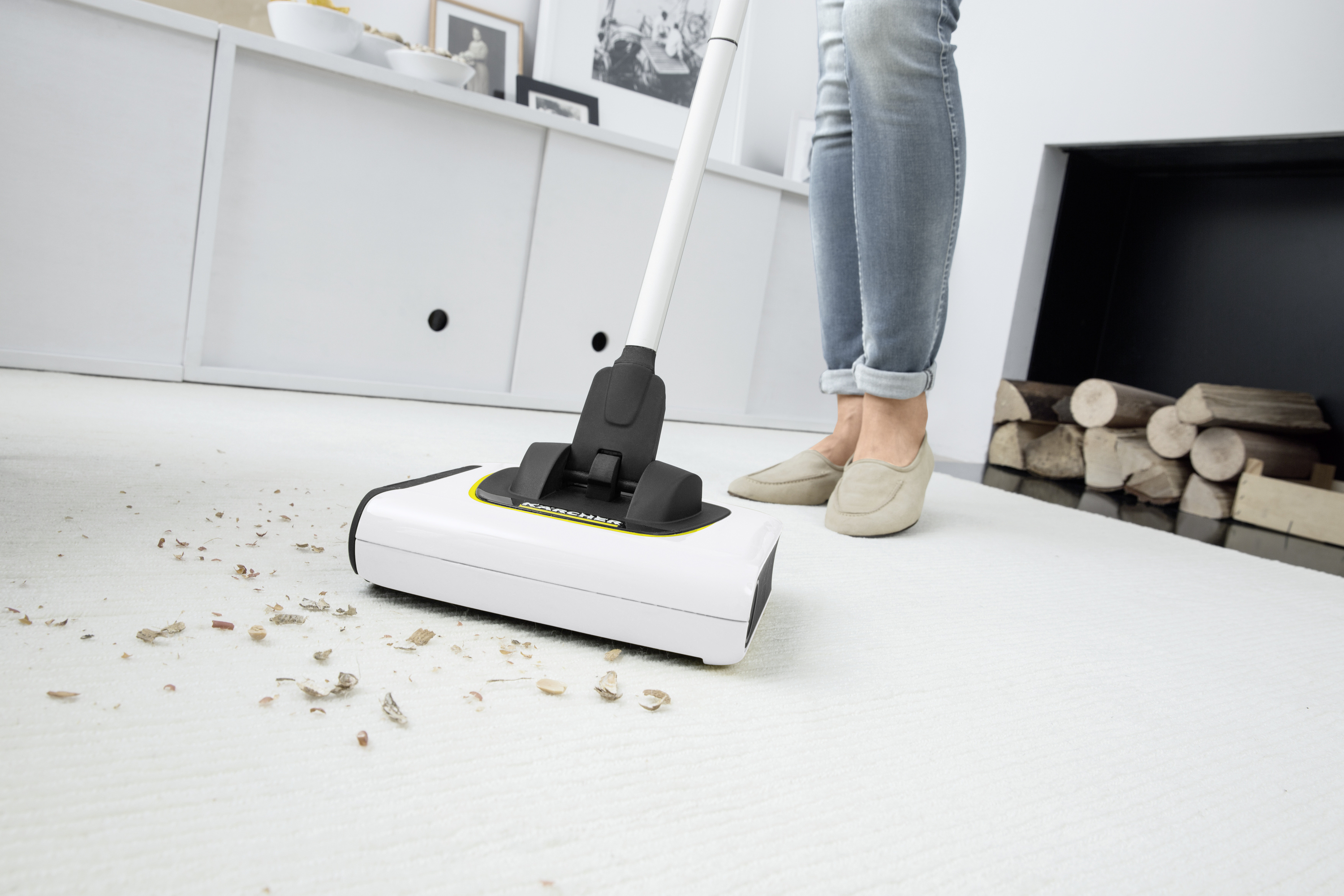A person in jeans and beige shoes is vacuuming crumbs from a white carpet in a living room. Logs can be seen in the background.