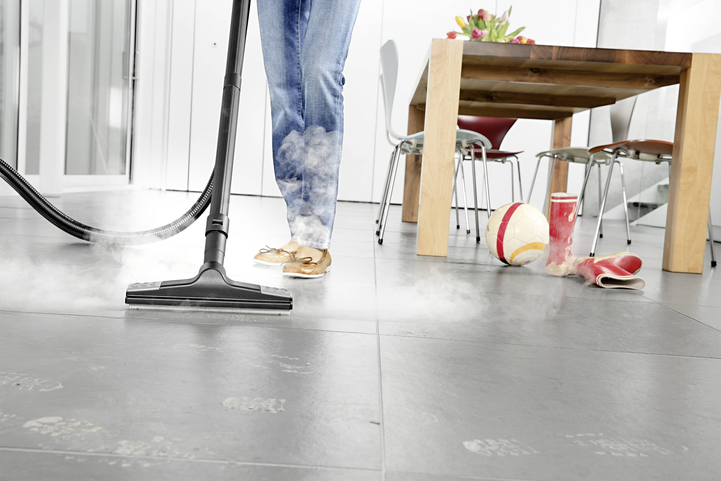 A person is cleaning a tiled floor with a steam cleaner. In the background, there is a table with chairs, a ball, and shoes.