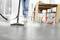 A person is cleaning a tiled floor with a steam cleaner. In the background, there is a table with chairs, a ball, and shoes.