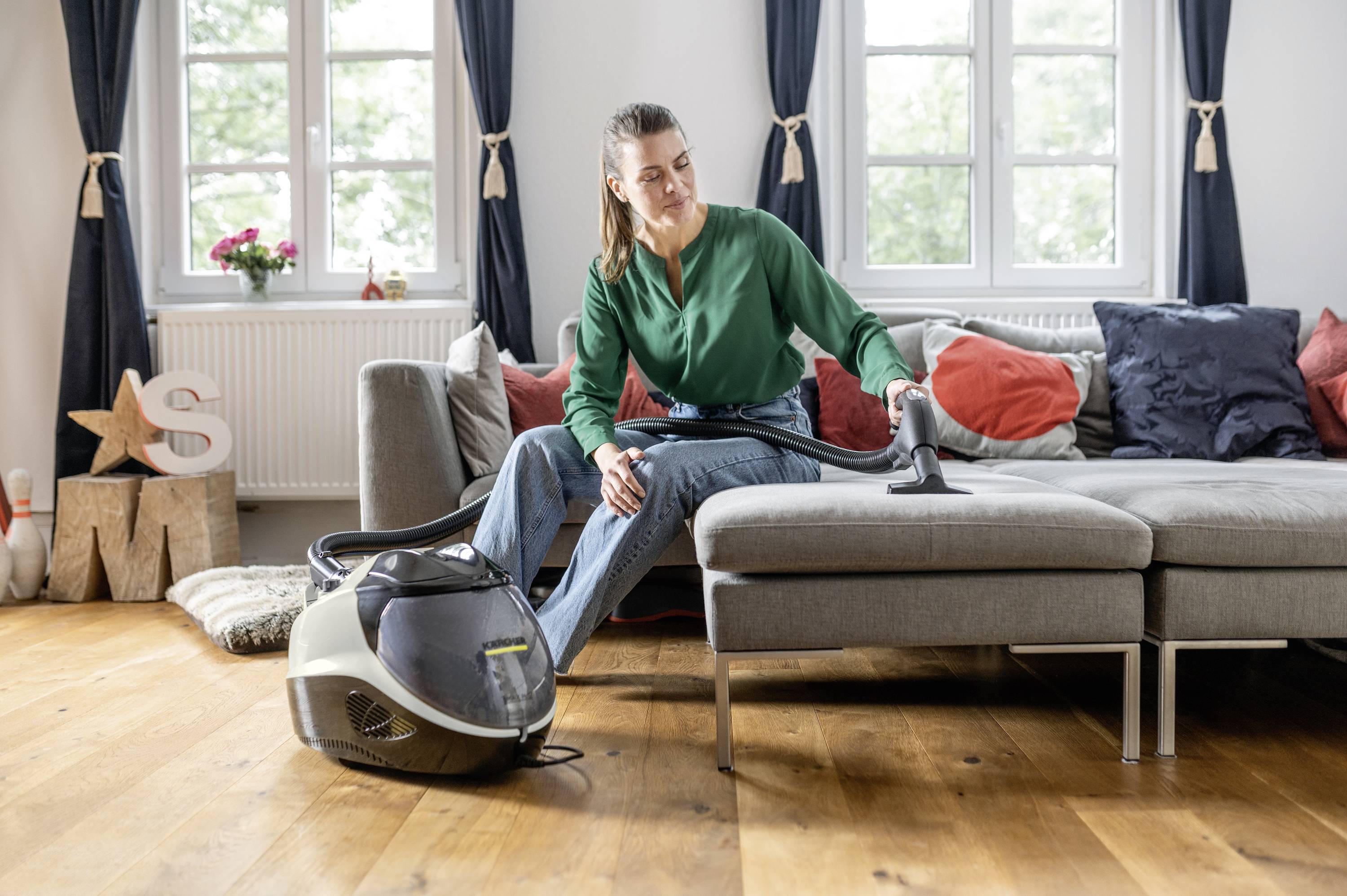 A woman in a green blouse is cleaning a sofa with a handheld vacuum cleaner. In the background, a large window and a grey sofa can be seen.