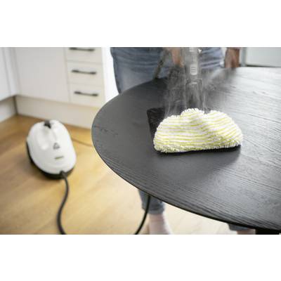 A person is cleaning a dark table with a steam cleaner. The steam cleaner is white and is standing on a wooden floor.