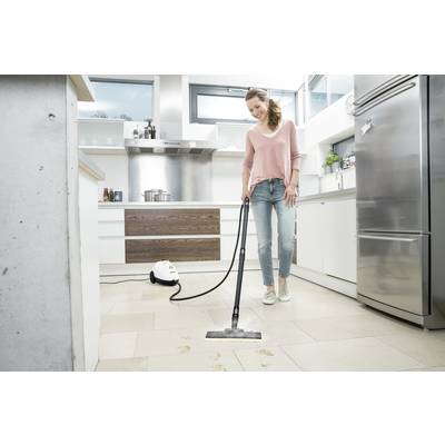 A woman is cleaning the kitchen floor with a steam cleaner. She is wearing a pink top and jeans. The kitchen looks modern and bright.