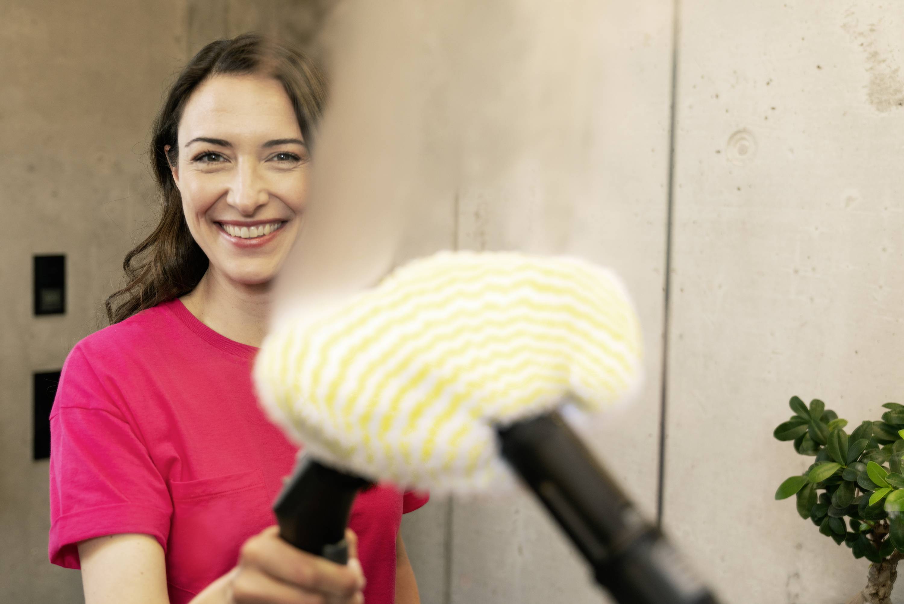 Woman in a red shirt smiling while holding a steam cleaner with a yellow head. Concrete background, plant on the right.