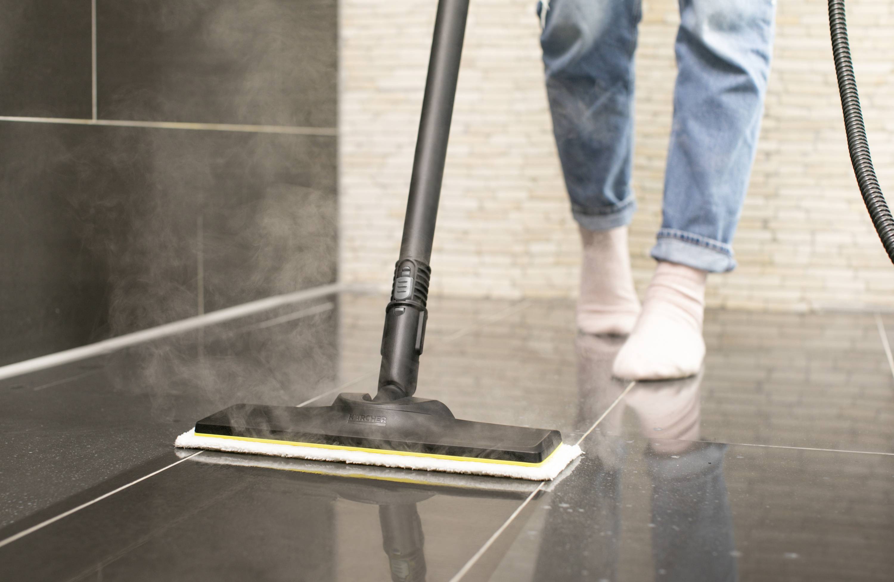 A person is cleaning shiny tiles with a steam cleaner in a room. The person is wearing jeans and socks.
