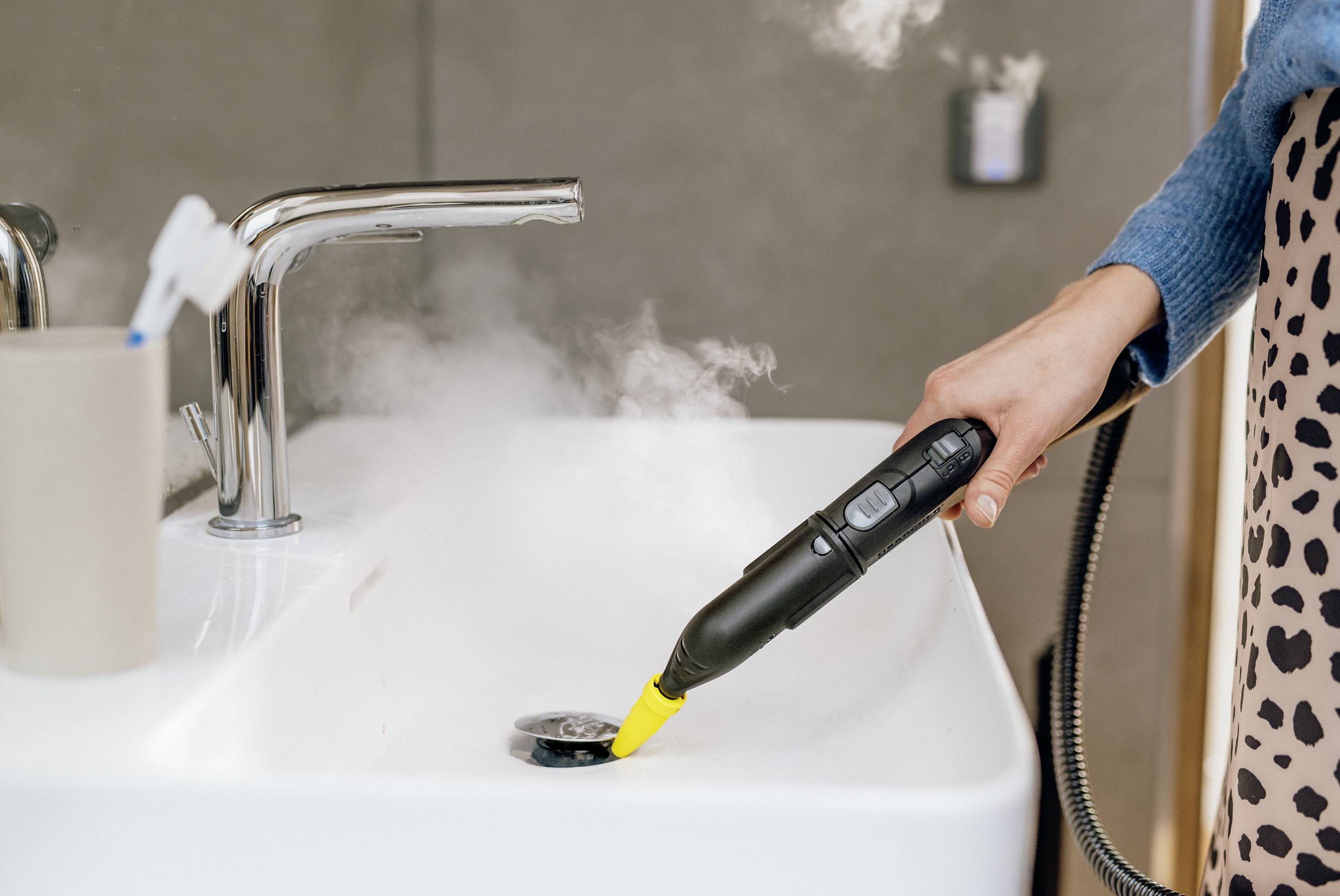 A person is cleaning the sink with a steam cleaner, with steam emerging from it. A tap and a toothbrush are in the background.