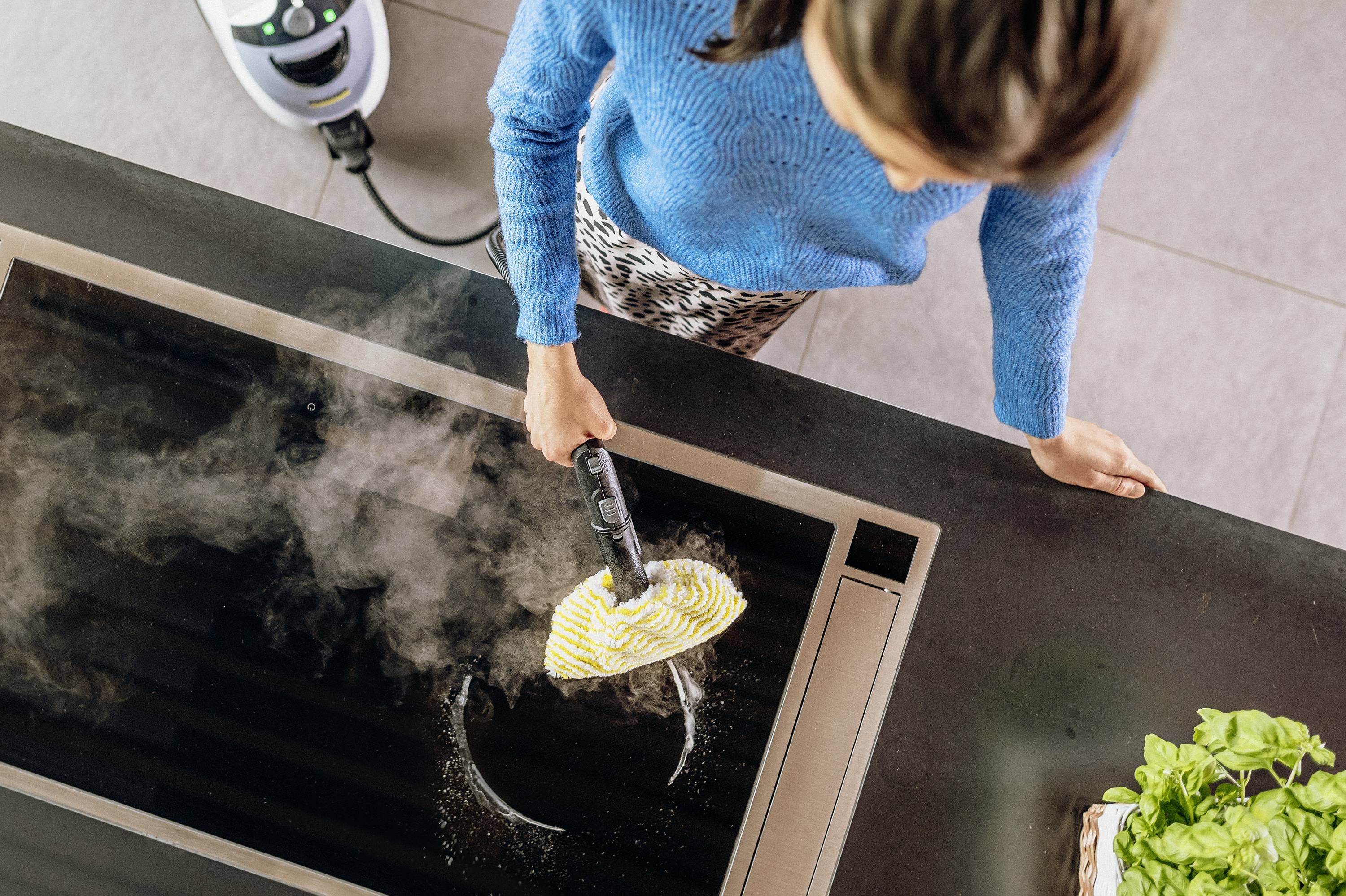 A person is cleaning a hob with a steam cleaner. The steam cleaning device is visible on the left side of the image. Fresh herbs are in the foreground.
