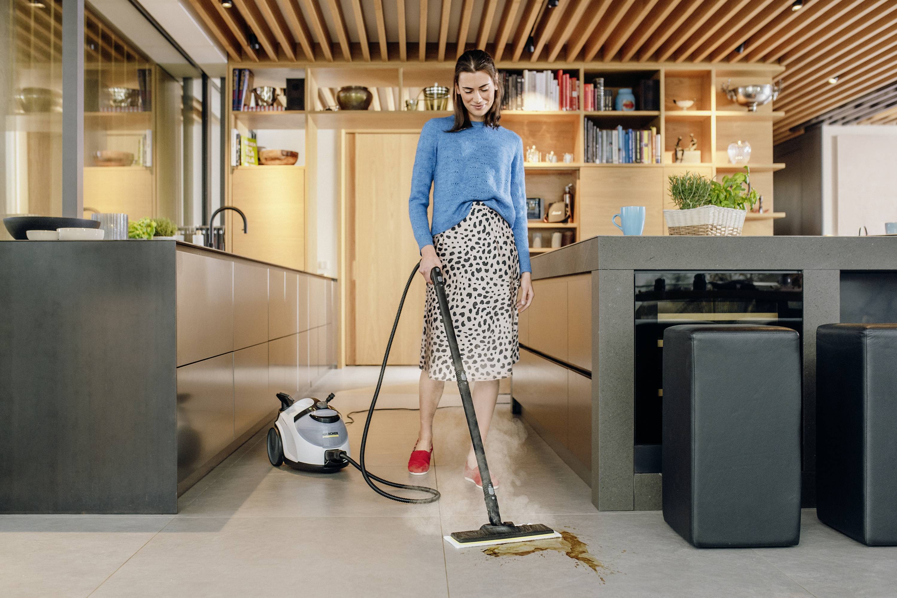A woman is mopping the kitchen floor with a steam cleaner. Shelves with books and kitchen utensils can be seen in the background.
