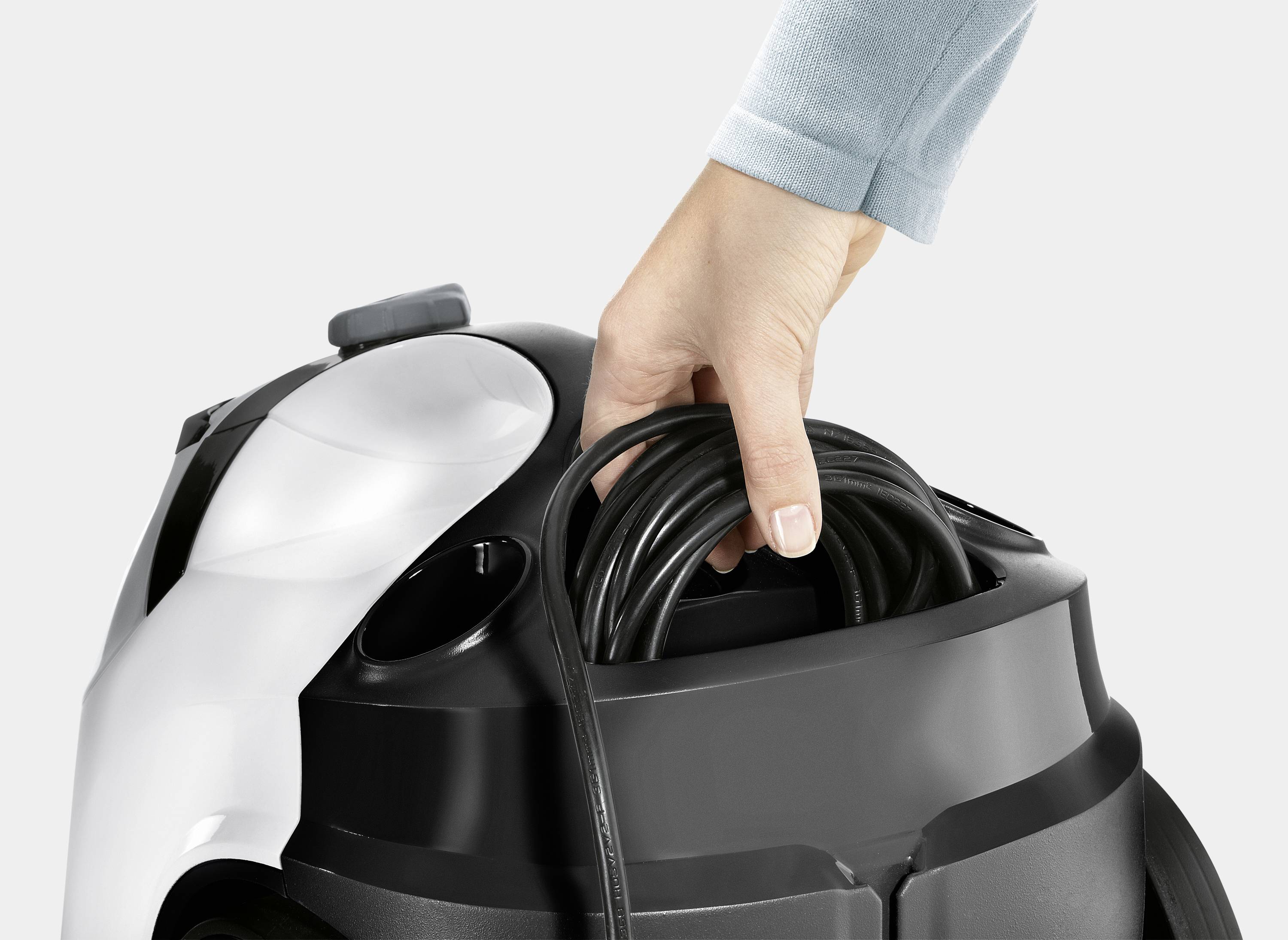 A hand places a black cable into the storage compartment of a black and white vacuum cleaner.