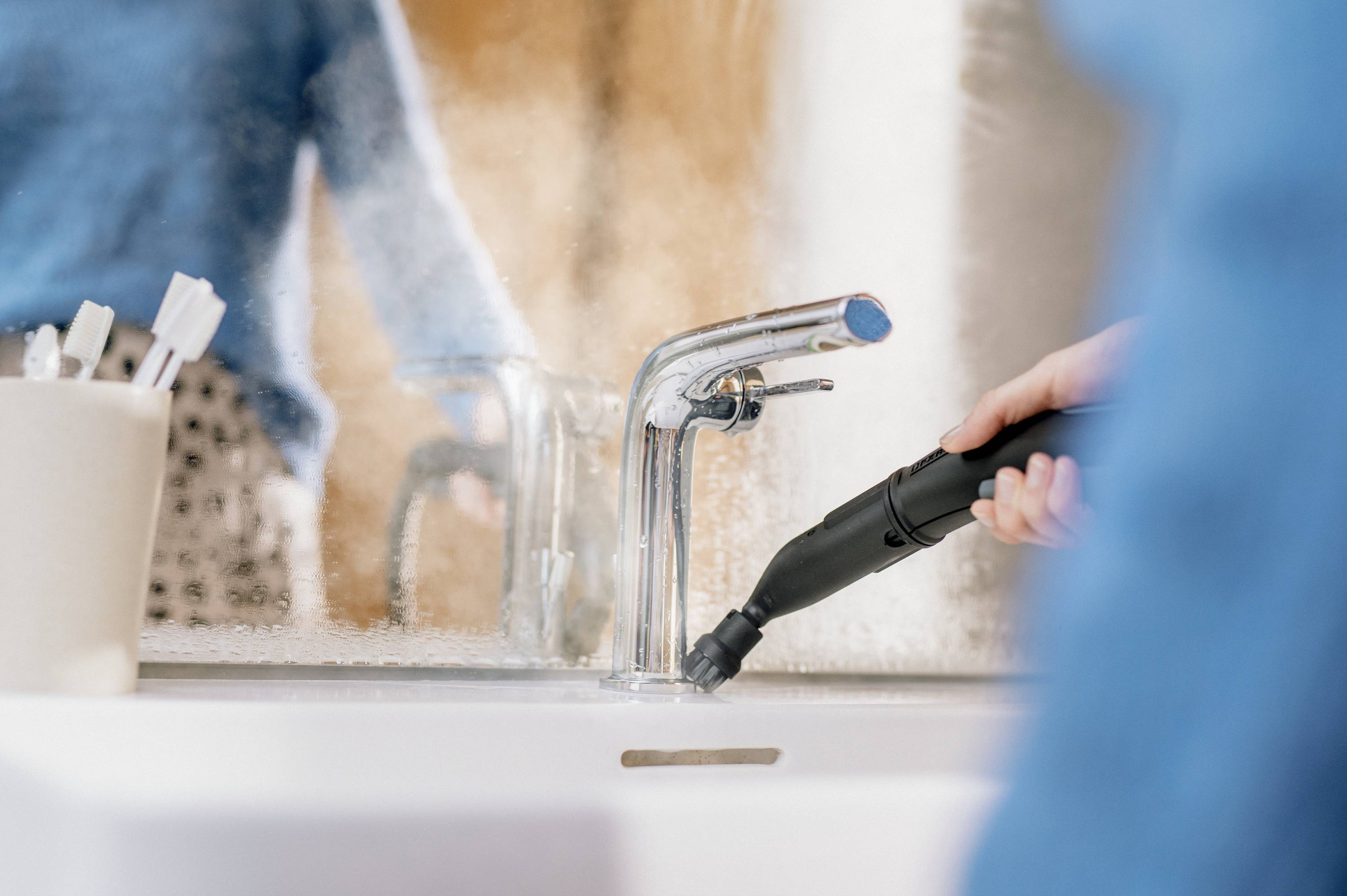A person is using a handheld steam cleaner to clean a tap. Blurred bathroom details are visible in the background.