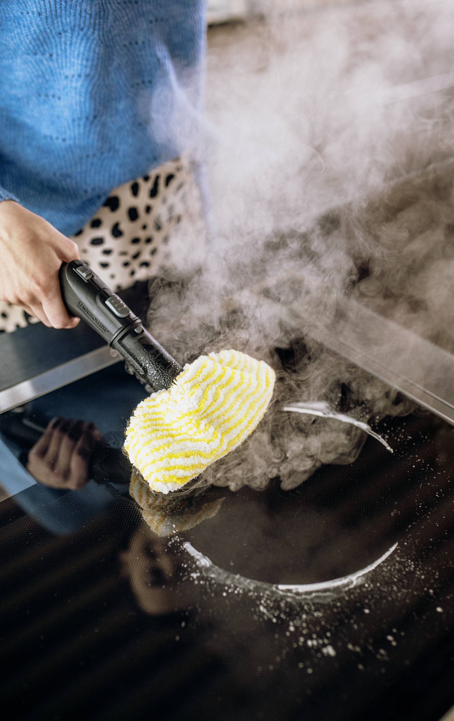 A person is cleaning a hob with a steam cleaner. Steam rises as the yellow brush moves across the glass surface.