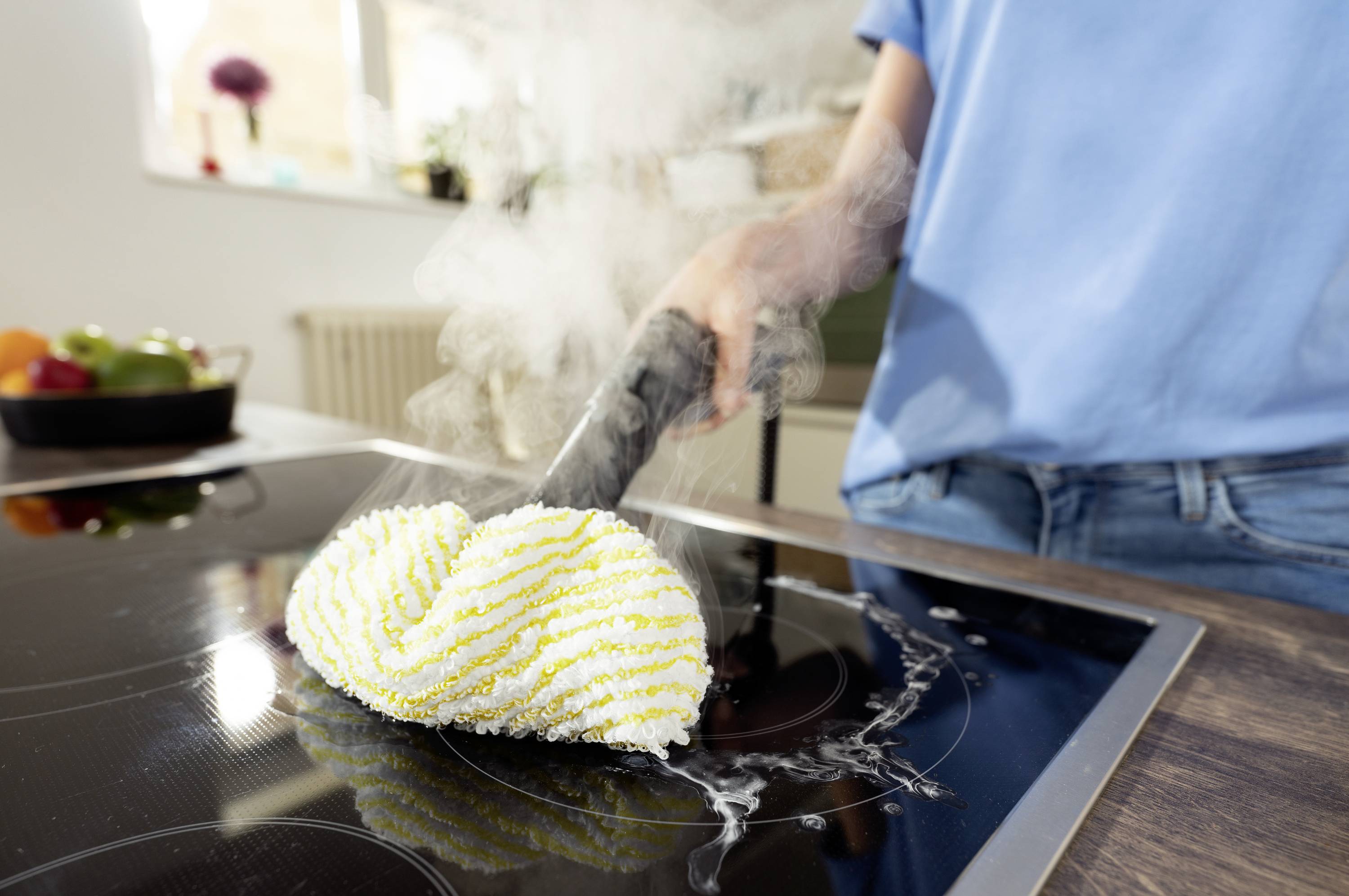 A person is cleaning a ceramic hob with a steam cleaner, with steam rising. Fruit is placed on a table in the background.