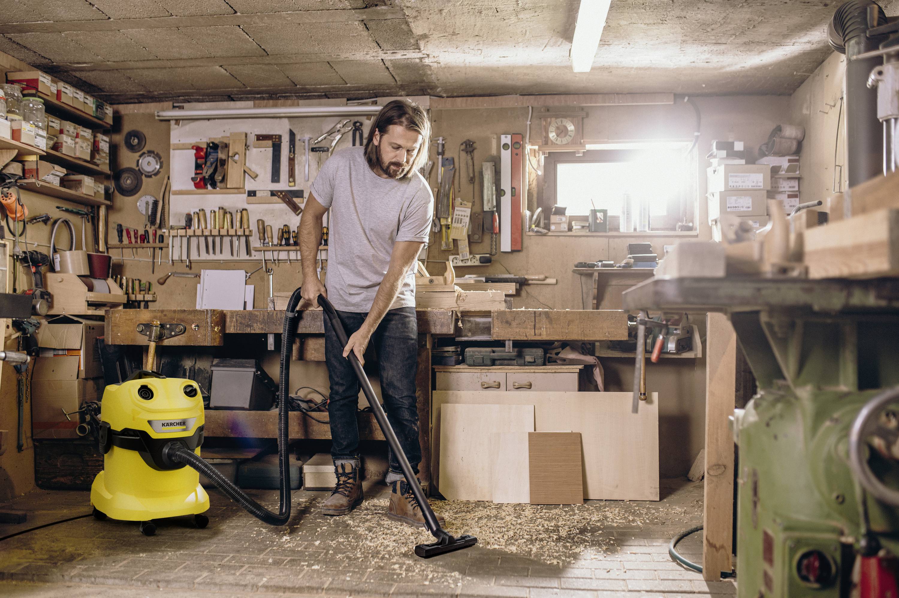 A person is vacuuming wood shavings in a workshop, surrounded by tools and a yellow vacuum cleaner.