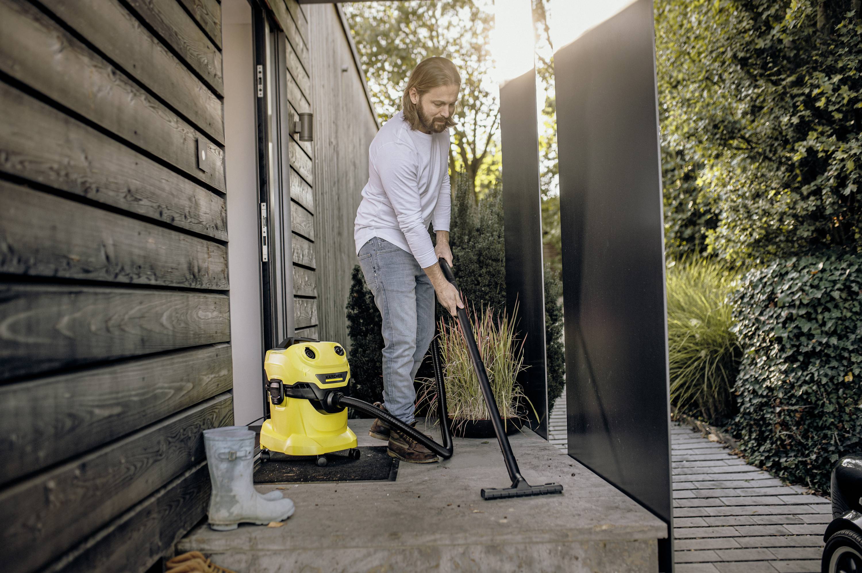 A man is cleaning the patio with a yellow vacuum cleaner on a sunny day. Wellington boots are standing next to the front door.