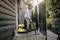 A man is cleaning the patio with a yellow vacuum cleaner on a sunny day. Wellington boots are standing next to the front door.