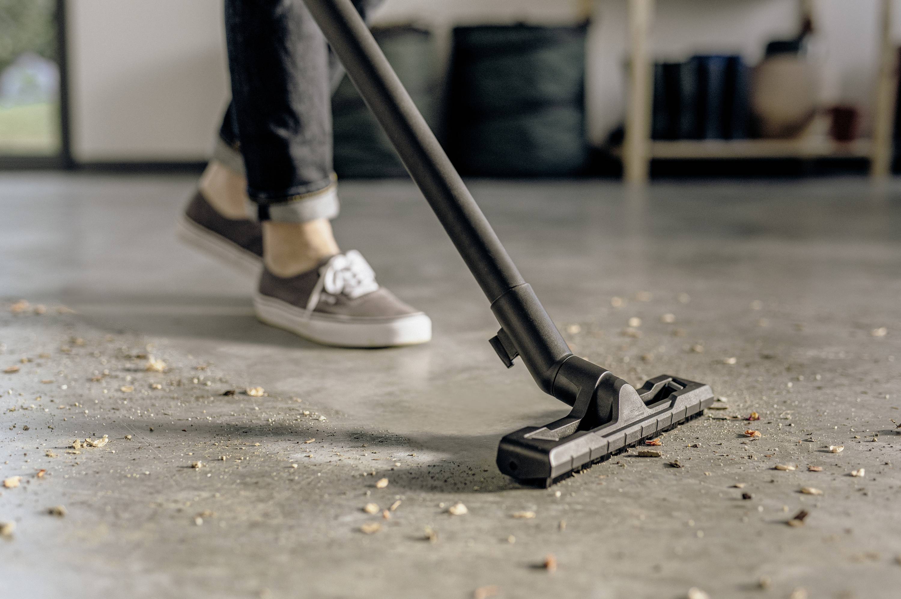 A person is hoovering a floor with scattered dirt, their legs in jeans and trainers visible. Background blurred.
