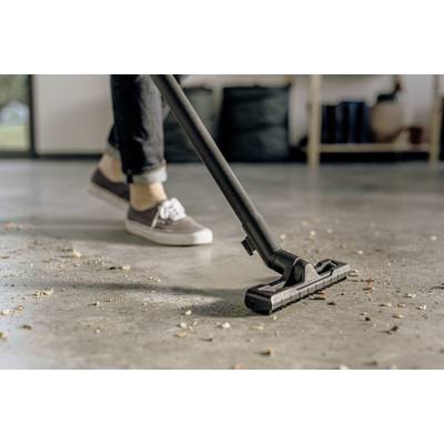 A person is hoovering a floor with scattered dirt, their legs in jeans and trainers visible. Background blurred.