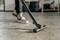 A person is hoovering a floor with scattered dirt, their legs in jeans and trainers visible. Background blurred.