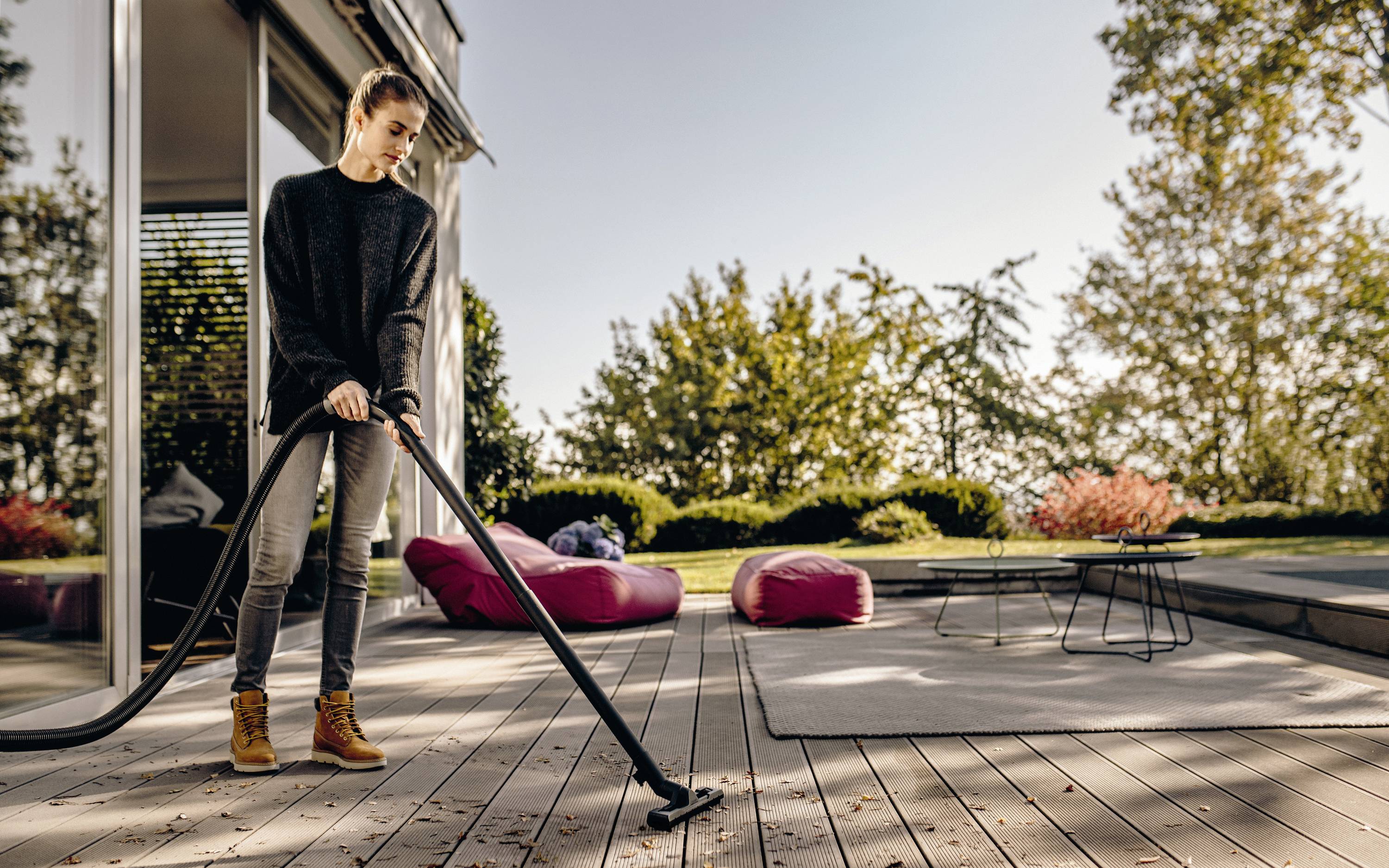 A person is vacuuming a patio, surrounded by garden furniture and trees in the background.