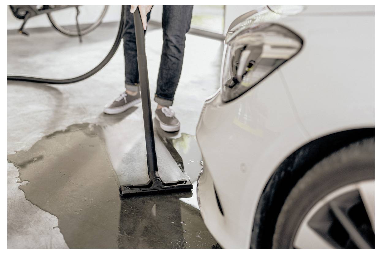 A person in jeans and sneakers uses a squeegee to clean water near a white car in a garage.