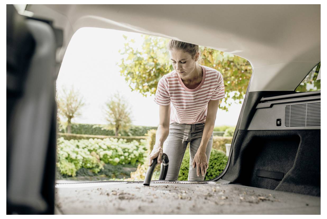 A person vacuuming the trunk of a car, wearing a striped shirt and jeans, with trees and shrubs visible in the background.