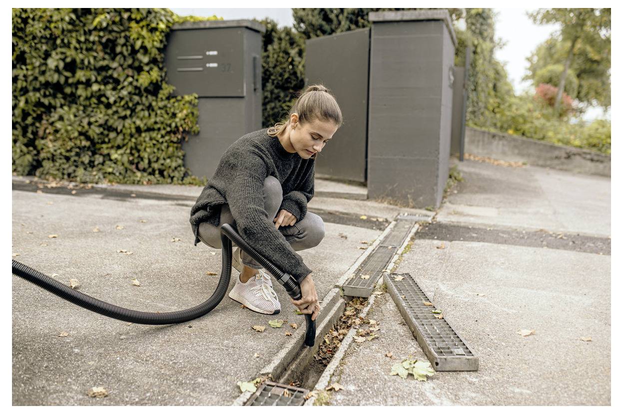 A person crouches on a driveway, cleaning debris from a drainage channel with a hose, surrounded by autumn leaves and greenery.