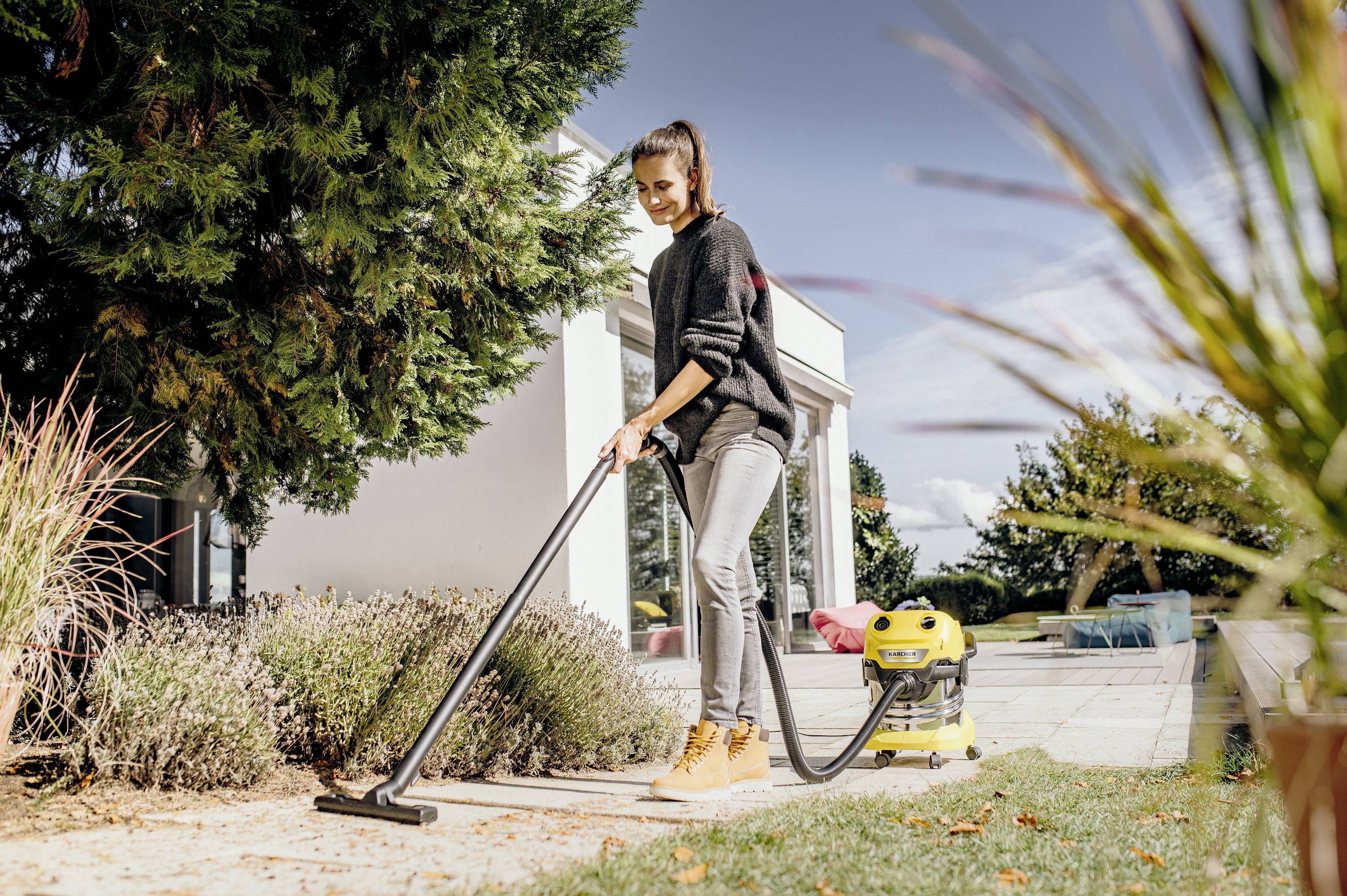 A woman is using a yellow industrial vacuum cleaner on a terrace to remove dirt and leaves. A modern house is visible in the background.