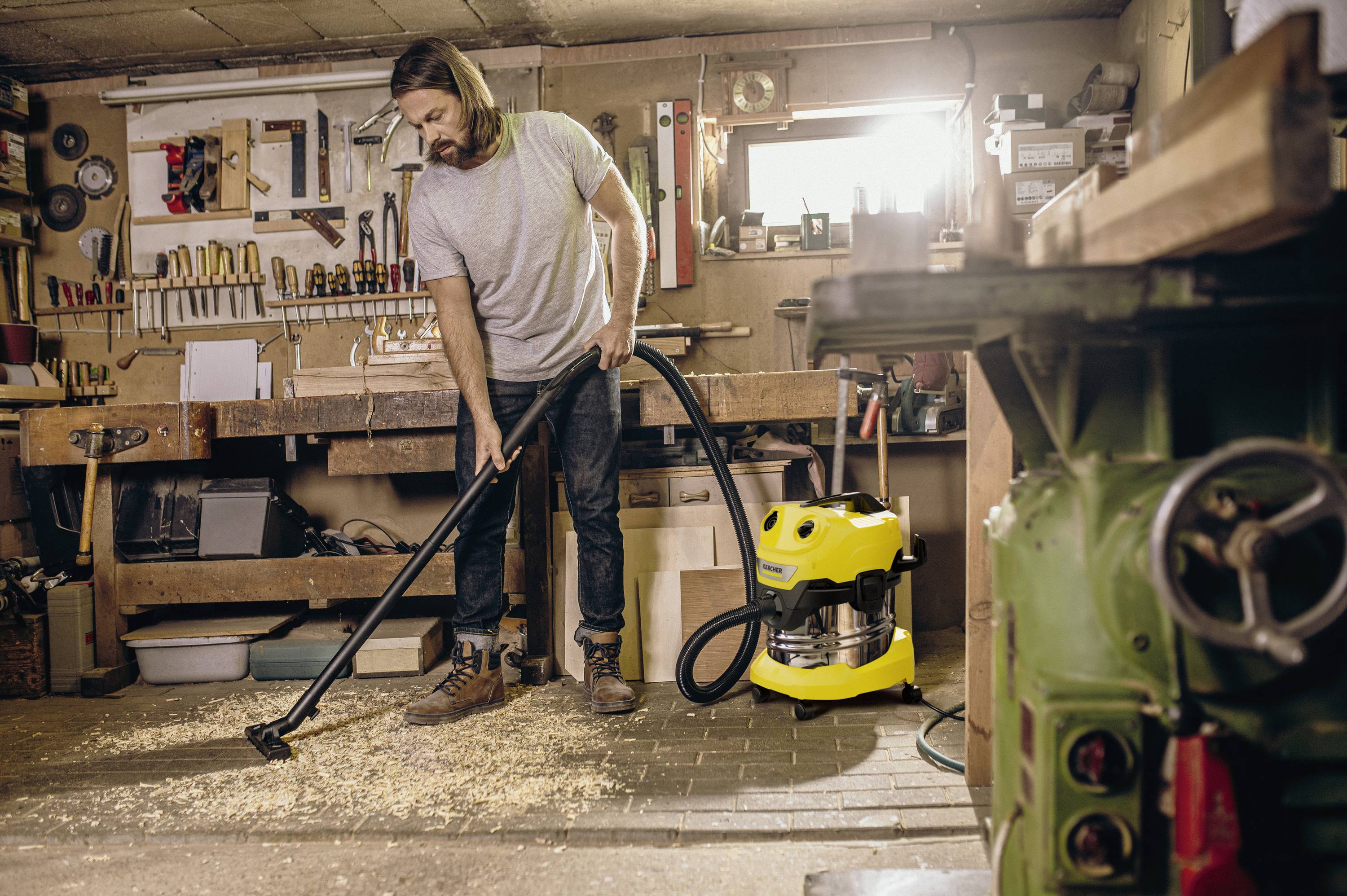 A man is cleaning the floor in a workshop with a vacuum cleaner. Many tools can be seen on the wall behind him.