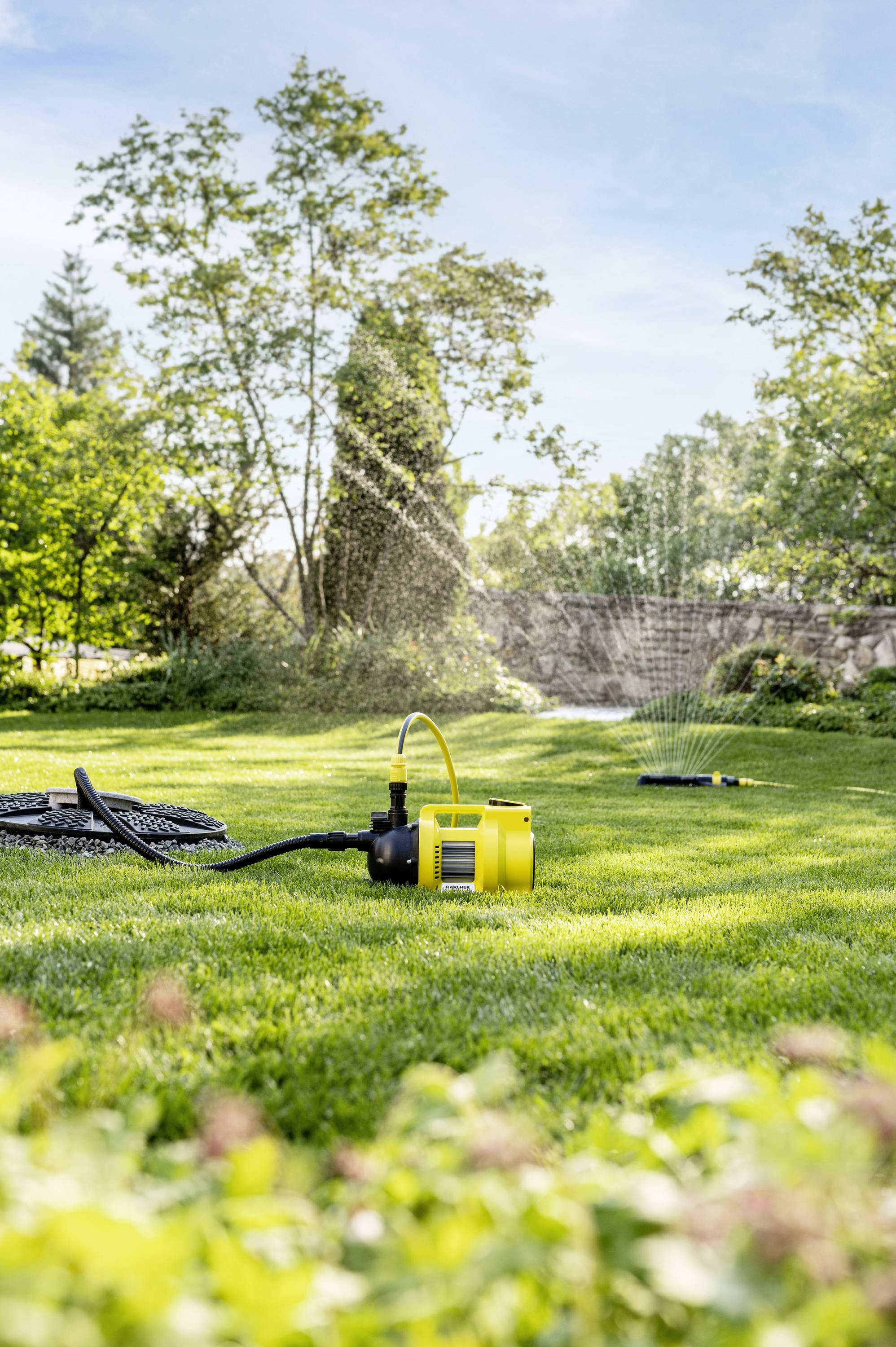 A yellow garden sprinkler is watering the lawn in a sunny garden, surrounded by trees and shrubs.