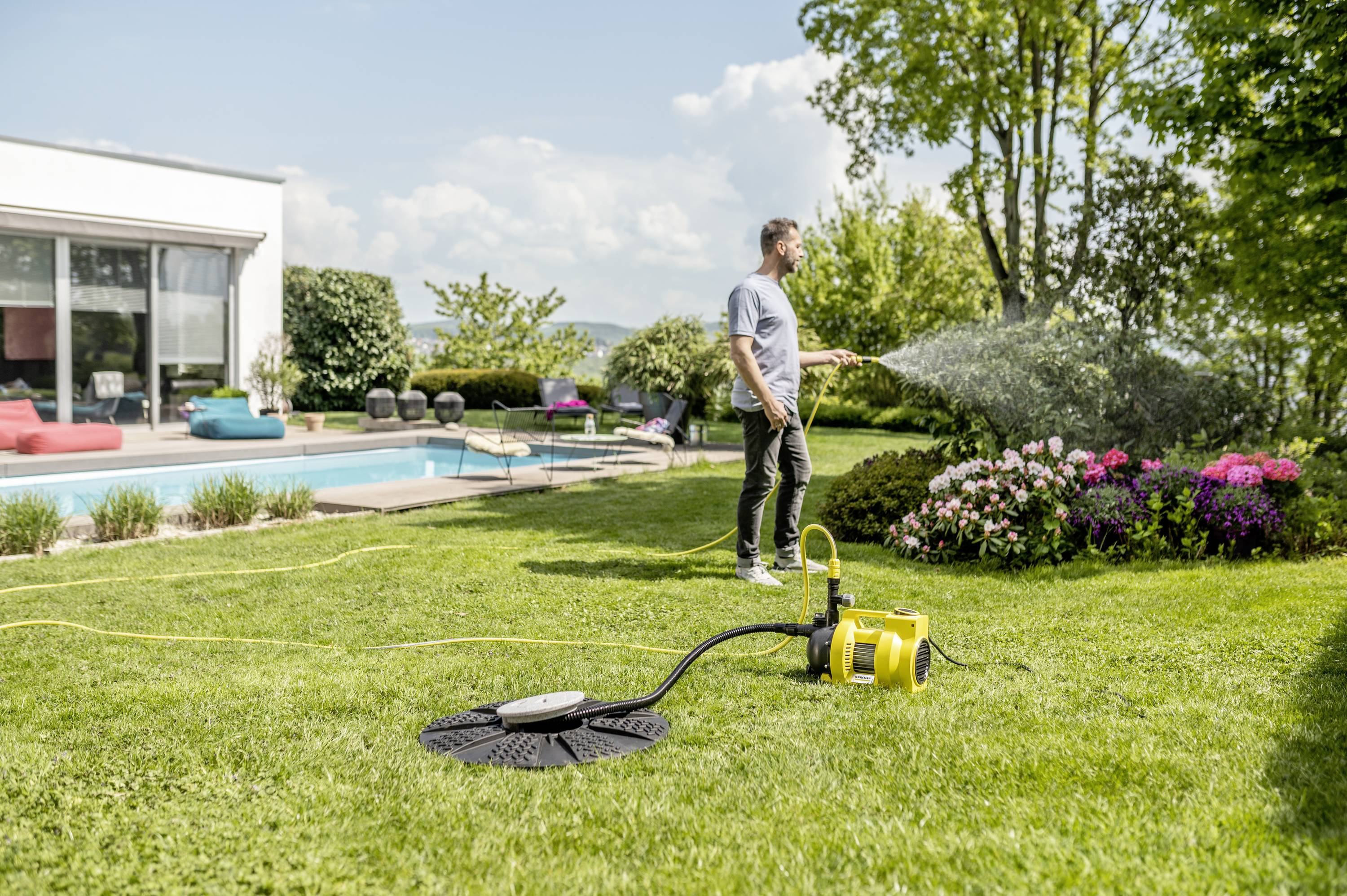 A man is watering plants with a garden hose in a garden next to a swimming pool. A modern house can be seen in the background.