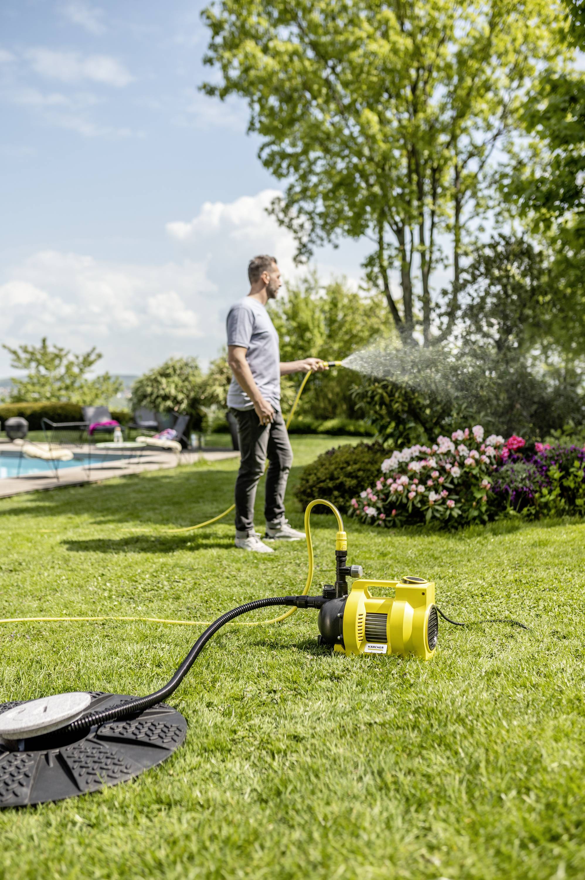 A man is watering plants with a garden hose in a sunny garden. A yellow water pump stands in the foreground.