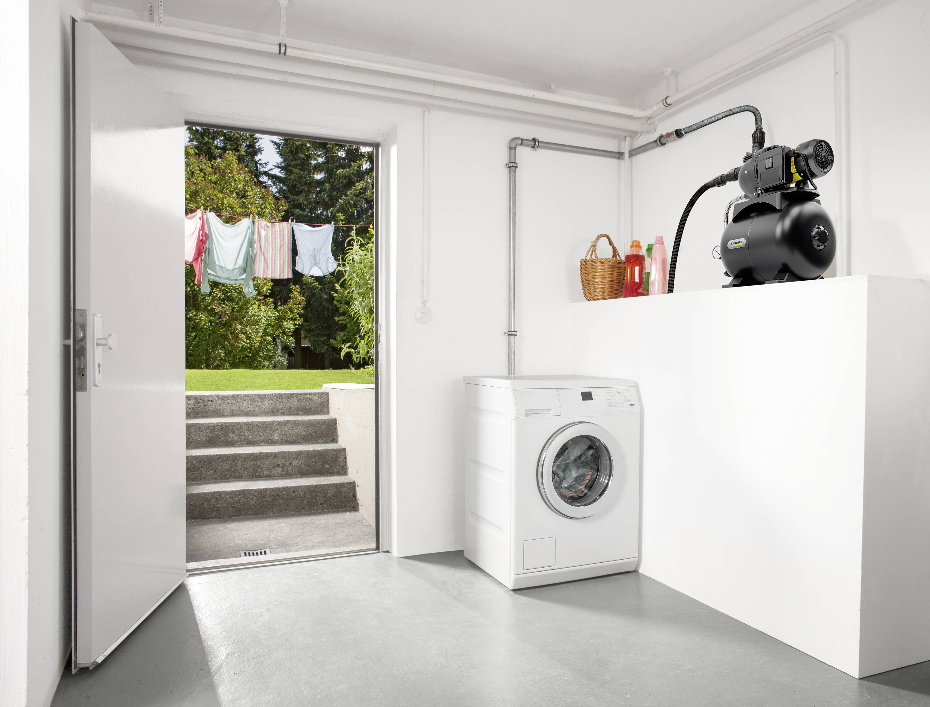 Utility room with a white washing machine, next to which sits a pump on a plinth. In the background, a door leads to a garden with a washing line.