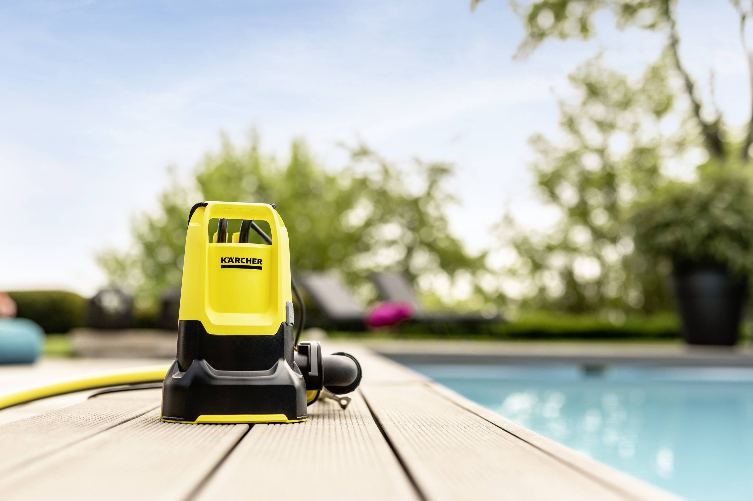 Yellow pressure washer on a wooden deck by the pool, background blurred with green trees and blue sky.