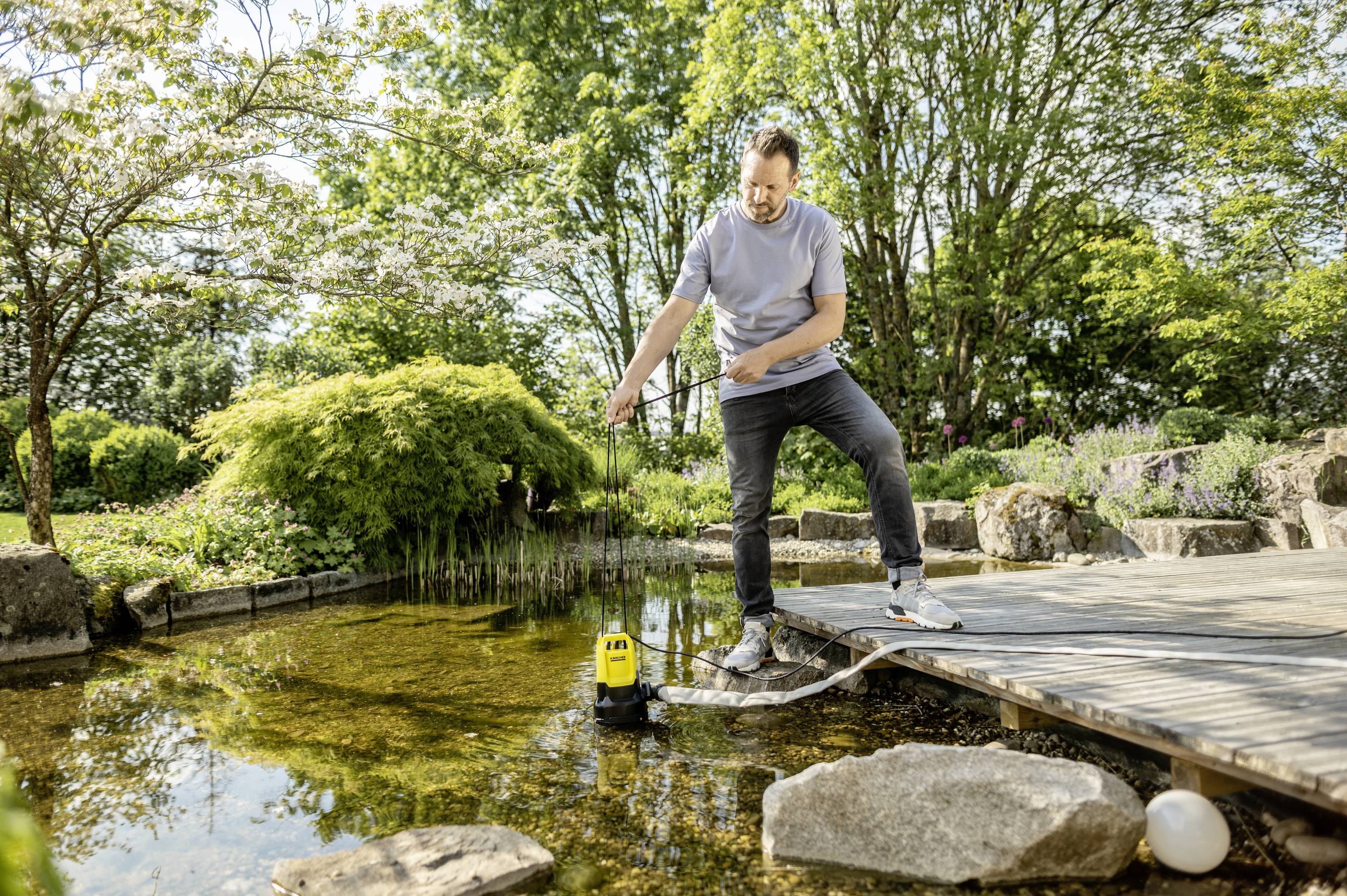 A man stands on a jetty by a pond, testing the water quality with a measuring device. In the background: lush vegetation.