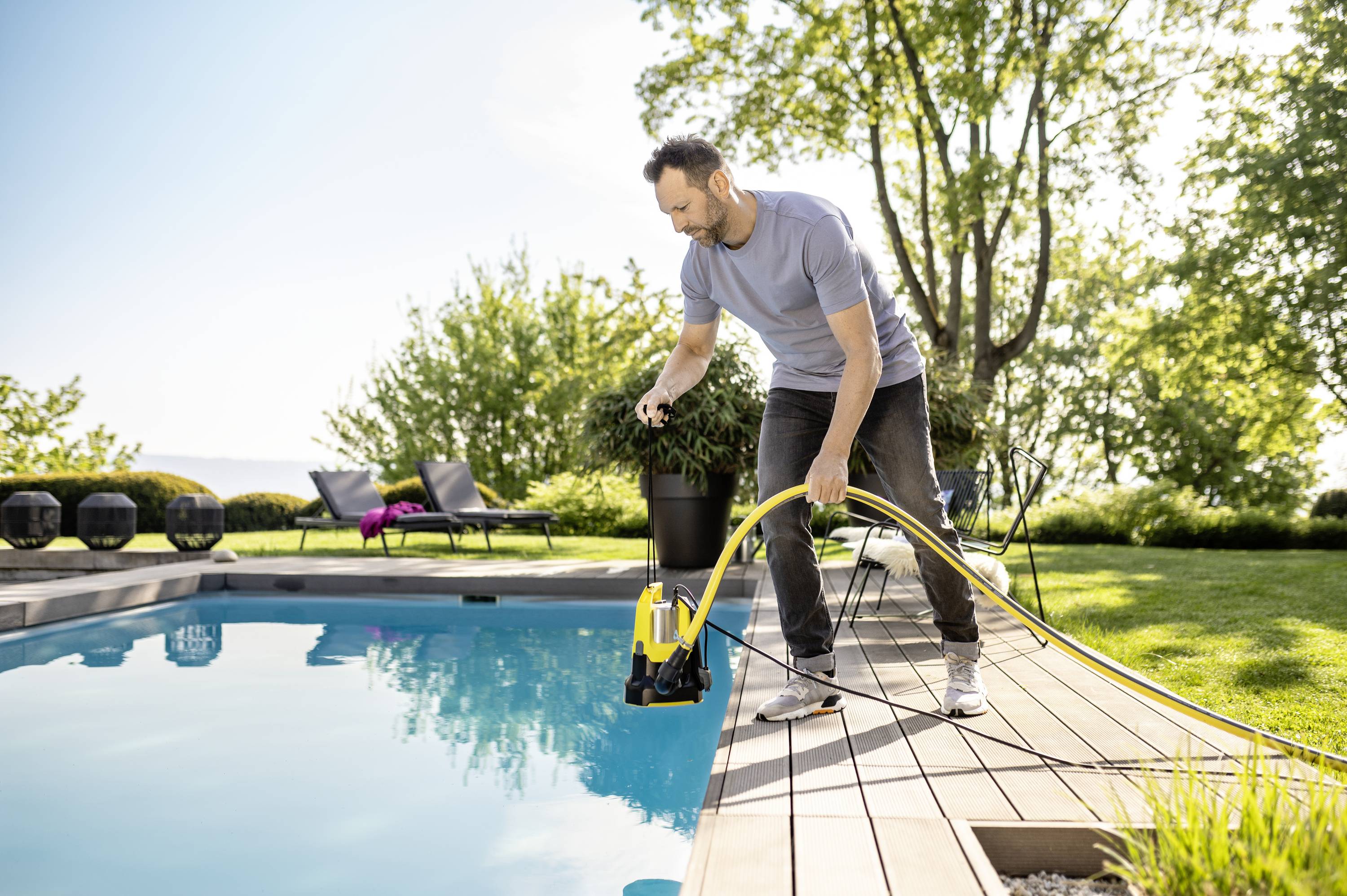 A man is cleaning a swimming pool with a submersible pump. Trees and garden chairs are visible in the background. It is a sunny day.