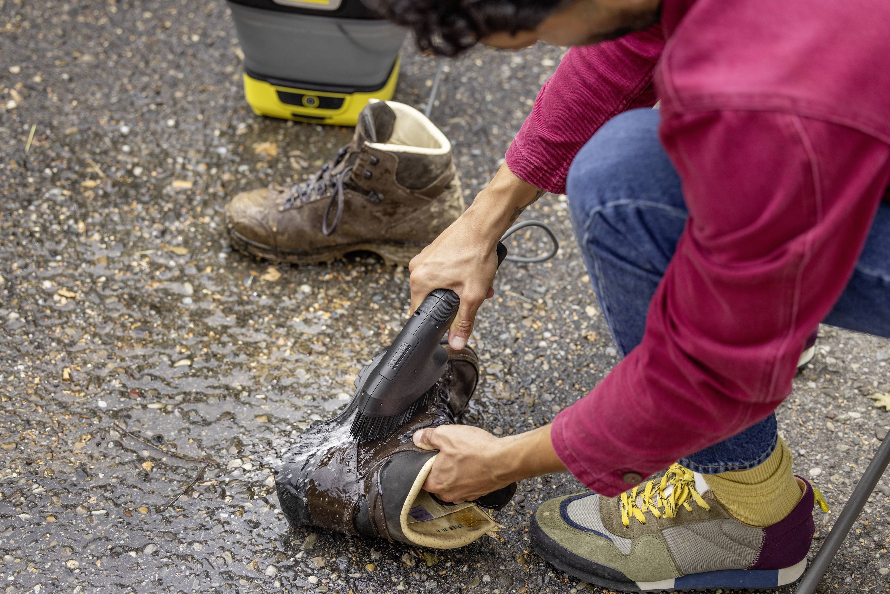 A person is cleaning work boots with a high-pressure washer on asphalt. Another already cleaned boot is standing beside it.
