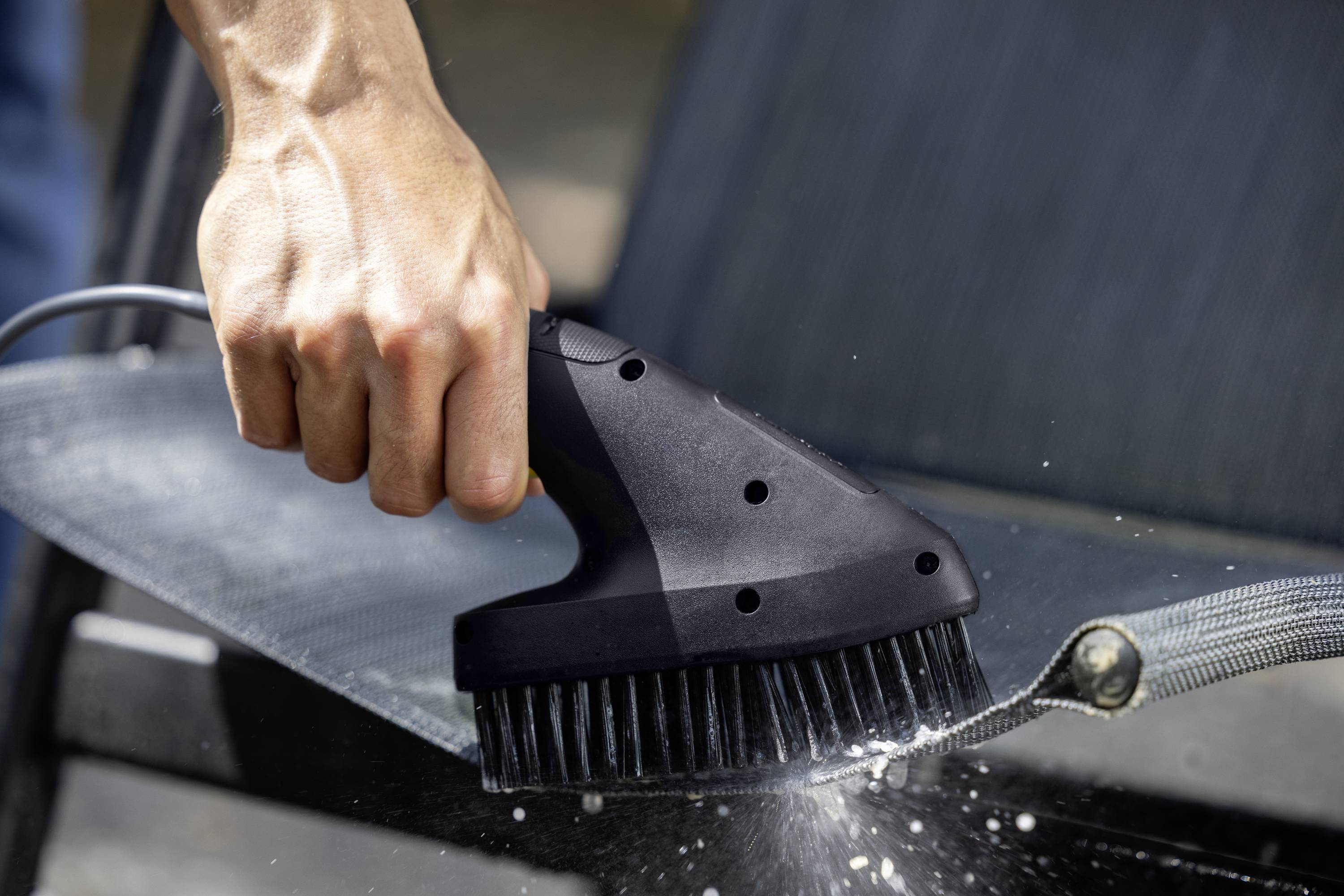 A person is cleaning the fabric of a garden chair with an electric brush. Water is splashing from the brush.
