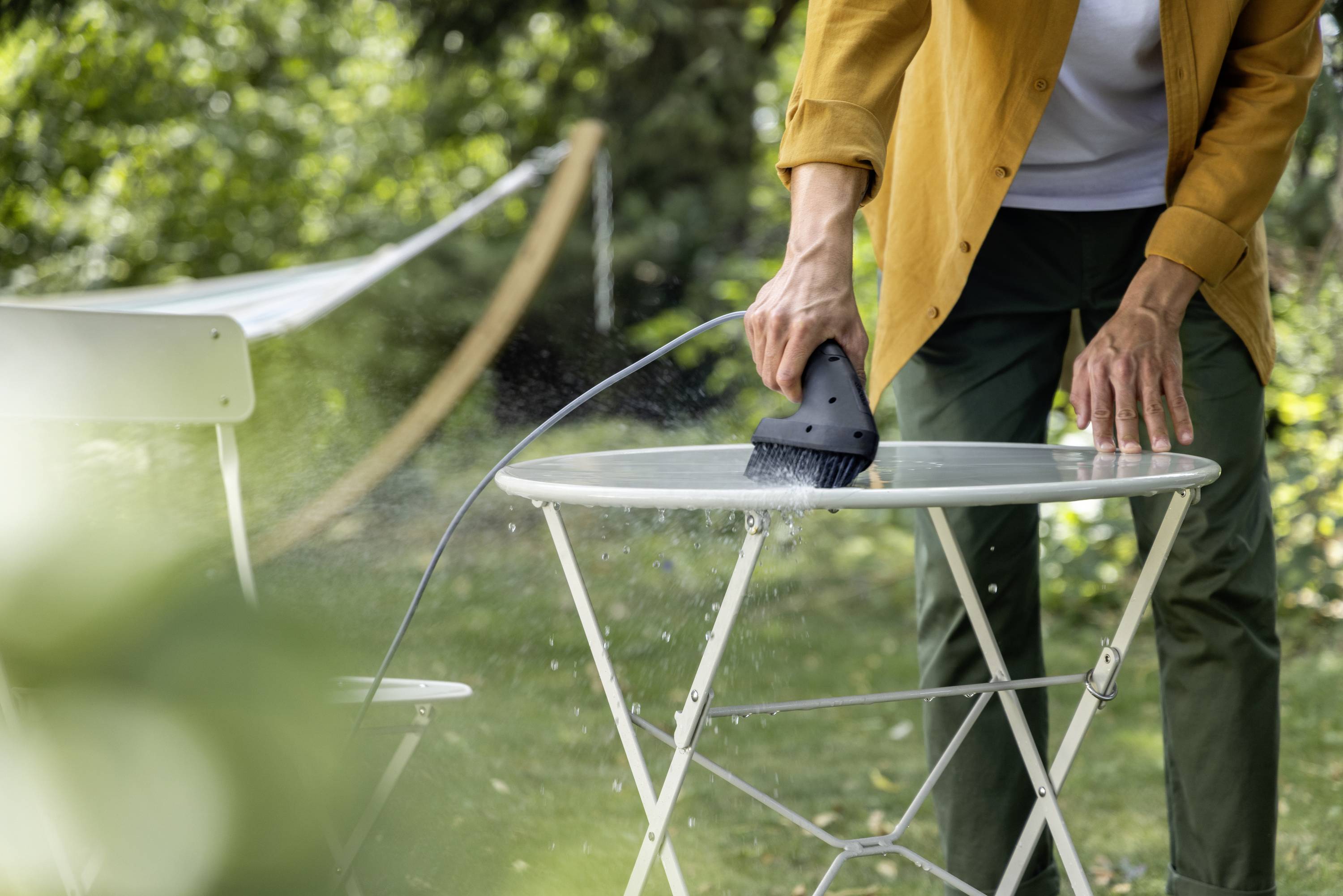 A person is cleaning a table outdoors with a handheld steam cleaner. In the background, a white chair and green vegetation can be seen.