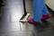A person is mopping a dark tiled floor with a floor cleaner. They are wearing blue trousers and pink slippers.