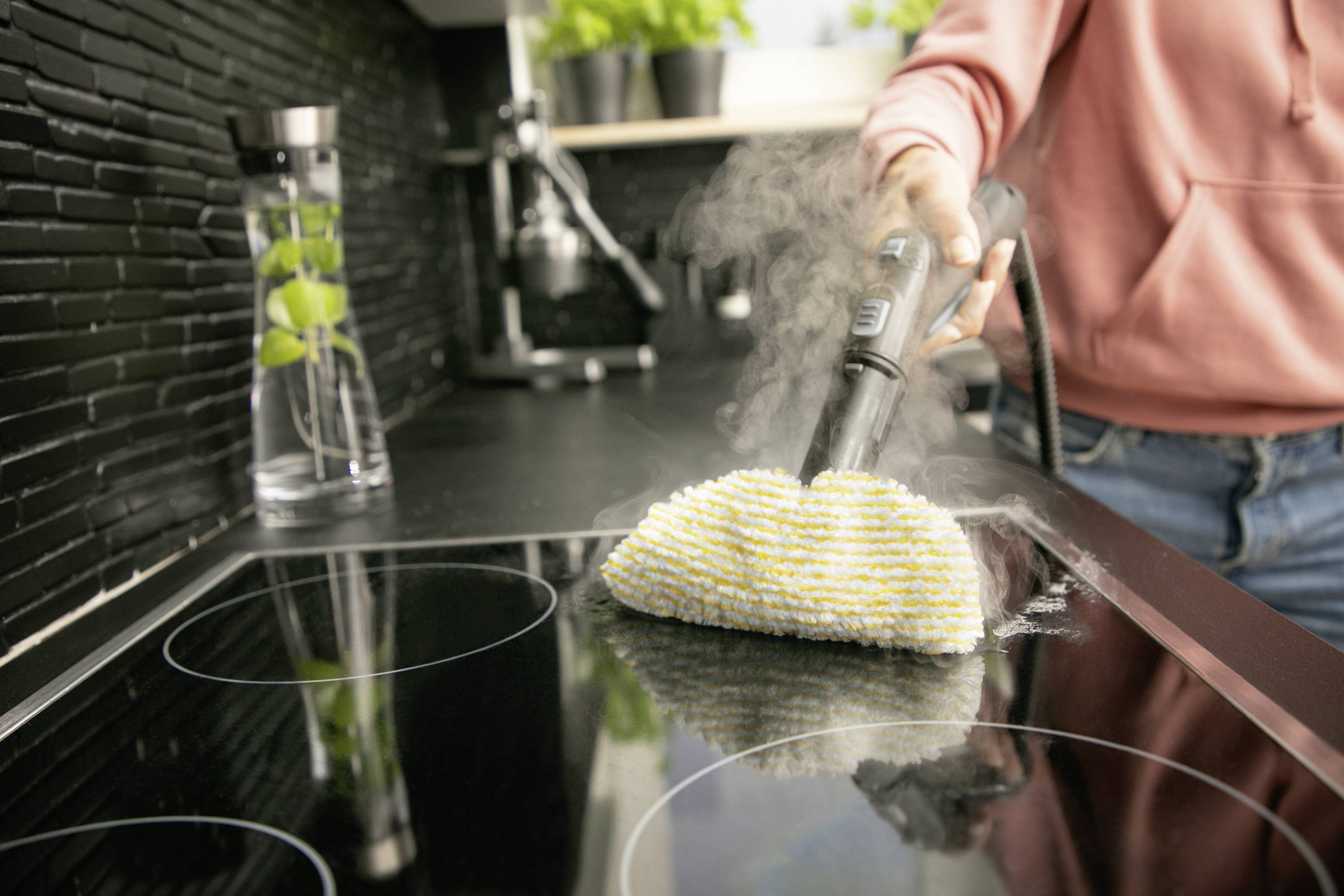 A person is cleaning a cooker with a steam cleaner, with steam rising. Kitchen utensils and plants are visible in the background.