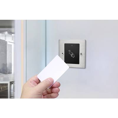 A hand is holding a card in front of a card reader. The reader is mounted on a wall and displays a symbol for contactless payments.