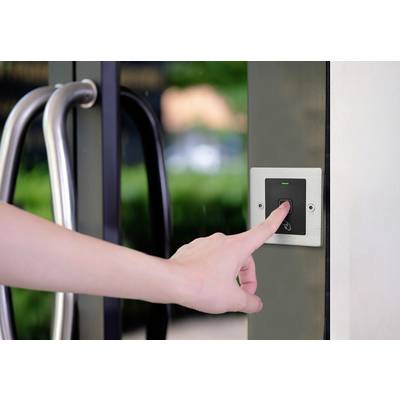 A hand presses a door access button next to a glass door, with a green indicator light illuminating. Blurred plants are visible in the background.