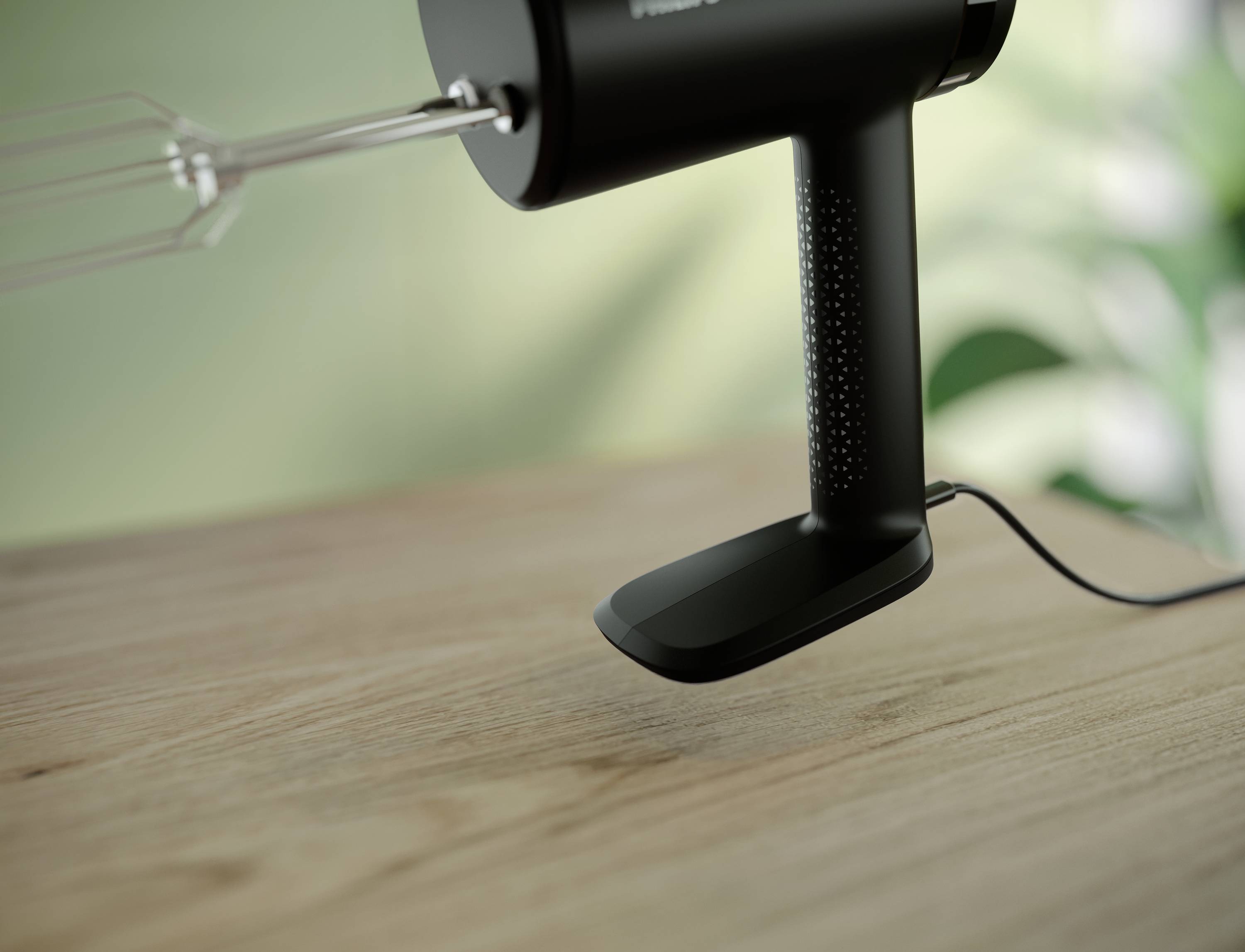 A black hand mixer sits on a wooden table; blurred plants are visible in the background.
