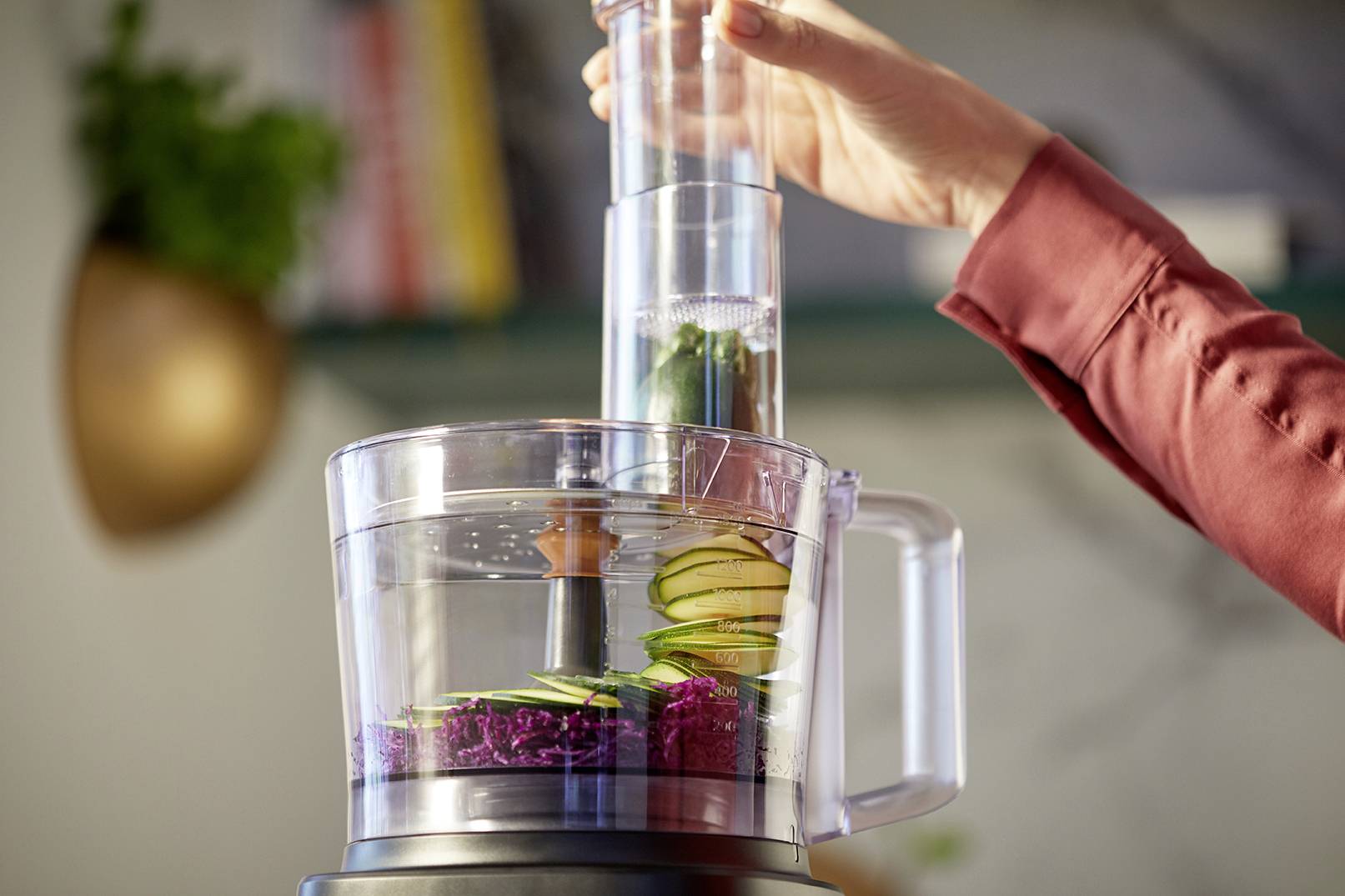 A hand operates a food processor, slicing thin vegetable pieces. Blurred plants and books are visible in the background.