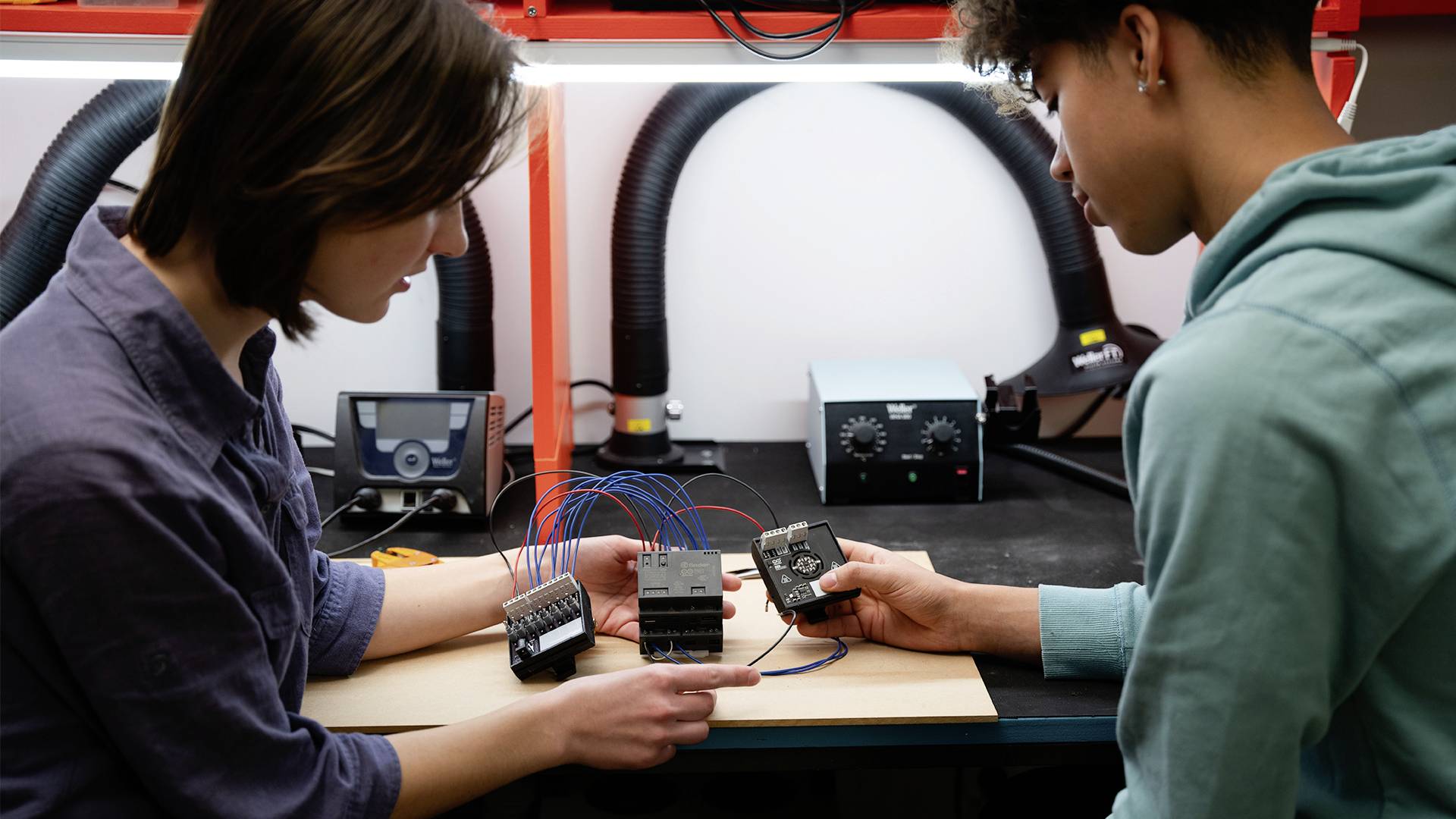 Two people are working on an electronic device with wires in a workshop. They are discussing and checking the connections.