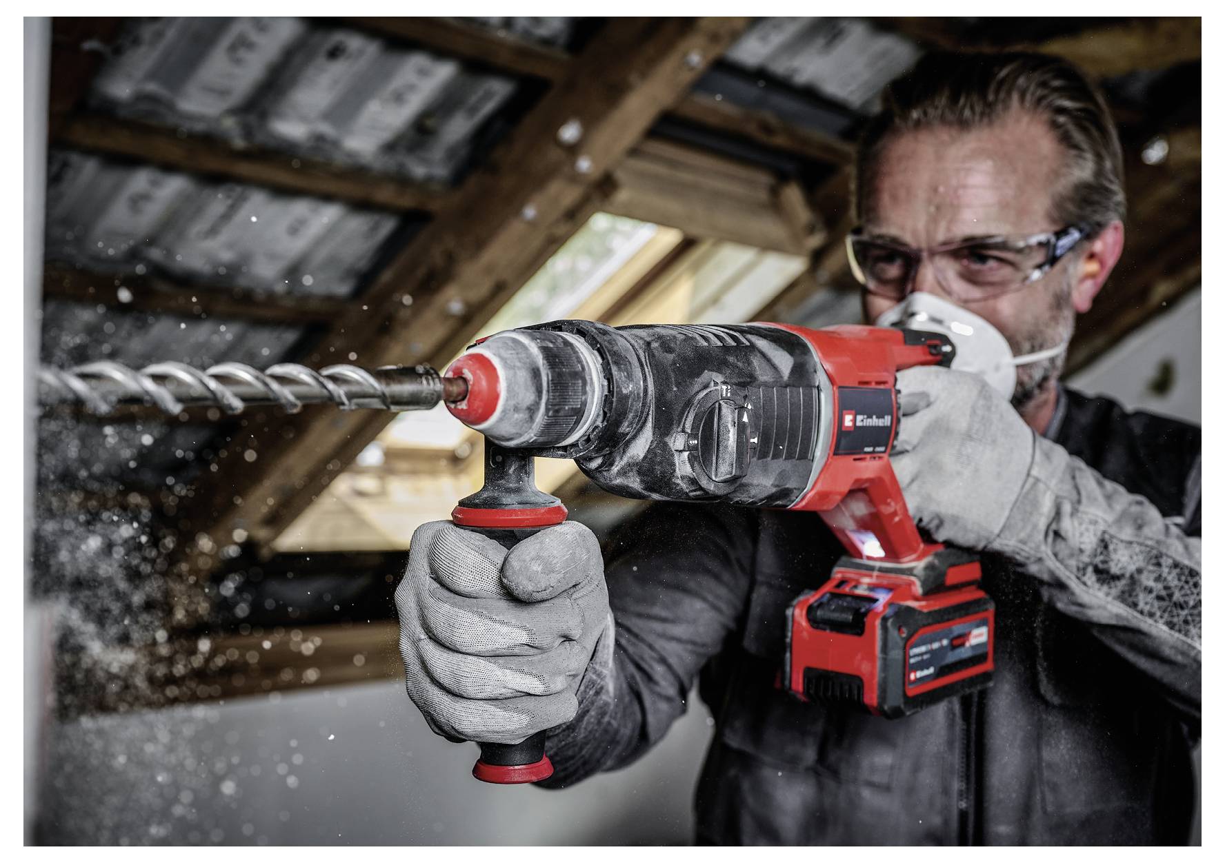 A person wearing safety goggles and gloves uses a red and black power drill to drill into a wooden beam inside a room under renovation.
