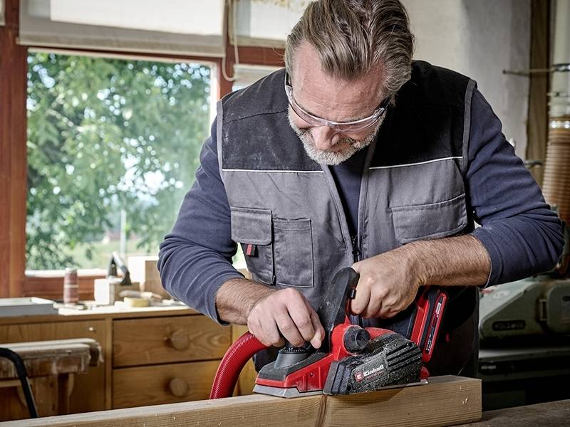 An older man is planing a wooden board in a workshop. He is wearing safety glasses and work attire. In the background, a window offers a view of trees.