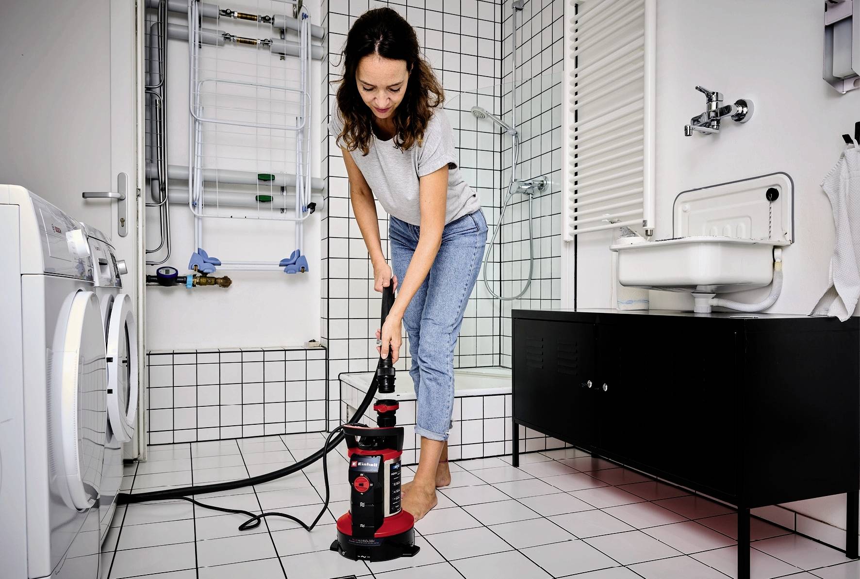 A woman is cleaning a white bathroom floor with a steam cleaner. Washing machine, sink, and radiator are in the background.